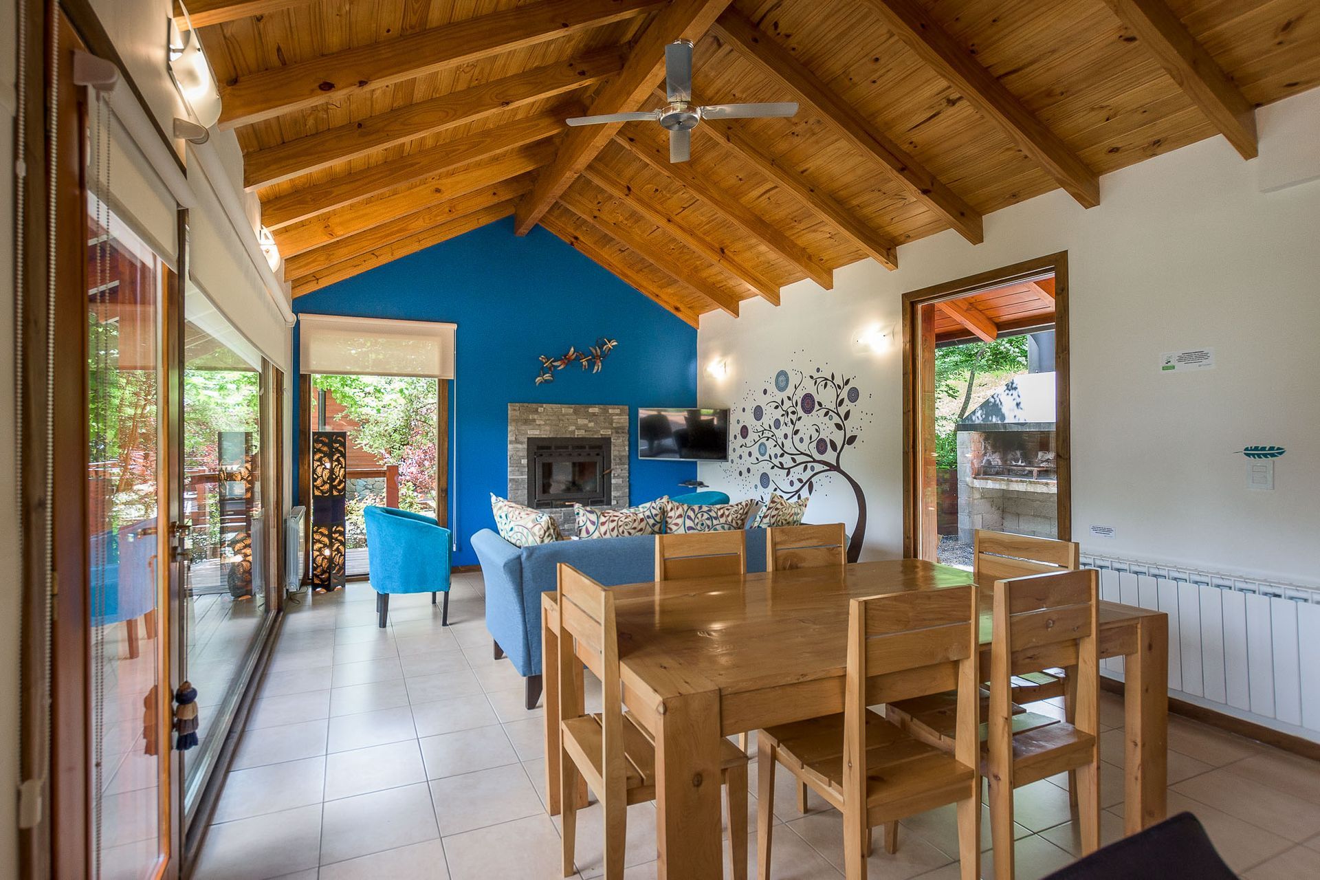 Un luminoso salón-comedor diáfano con techo abovedado de vigas de madera, una pared decorativa azul y suelos de baldosas.