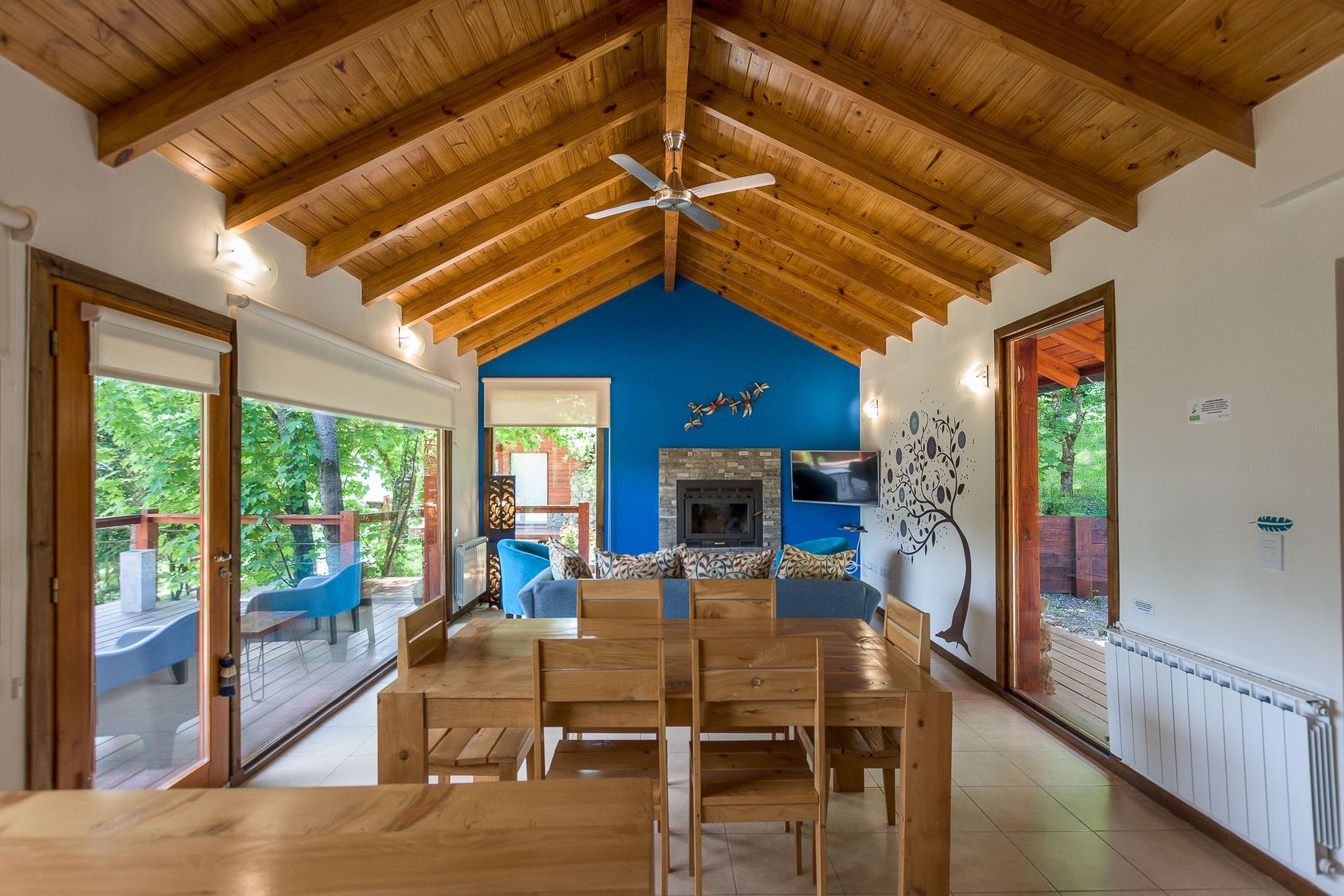 Una mesa de comedor de madera se encuentra en la sala de estar de una cabaña con techo abovedado de madera, una pared decorativa azul, chimenea y patio.