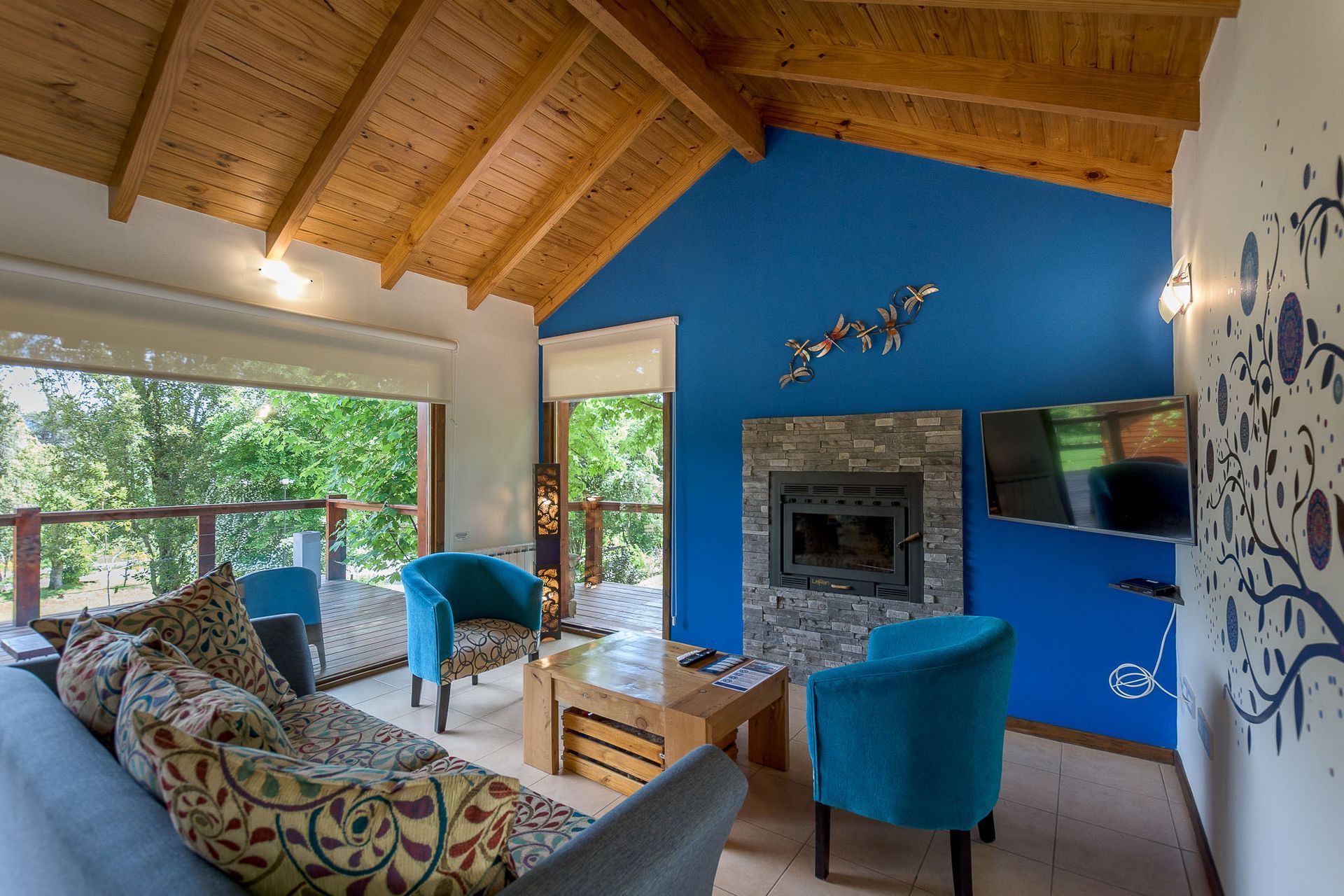Una acogedora sala de estar con vigas de madera, una pared decorativa de color azul vibrante, una chimenea de piedra, dos sillones azules y un sofá.