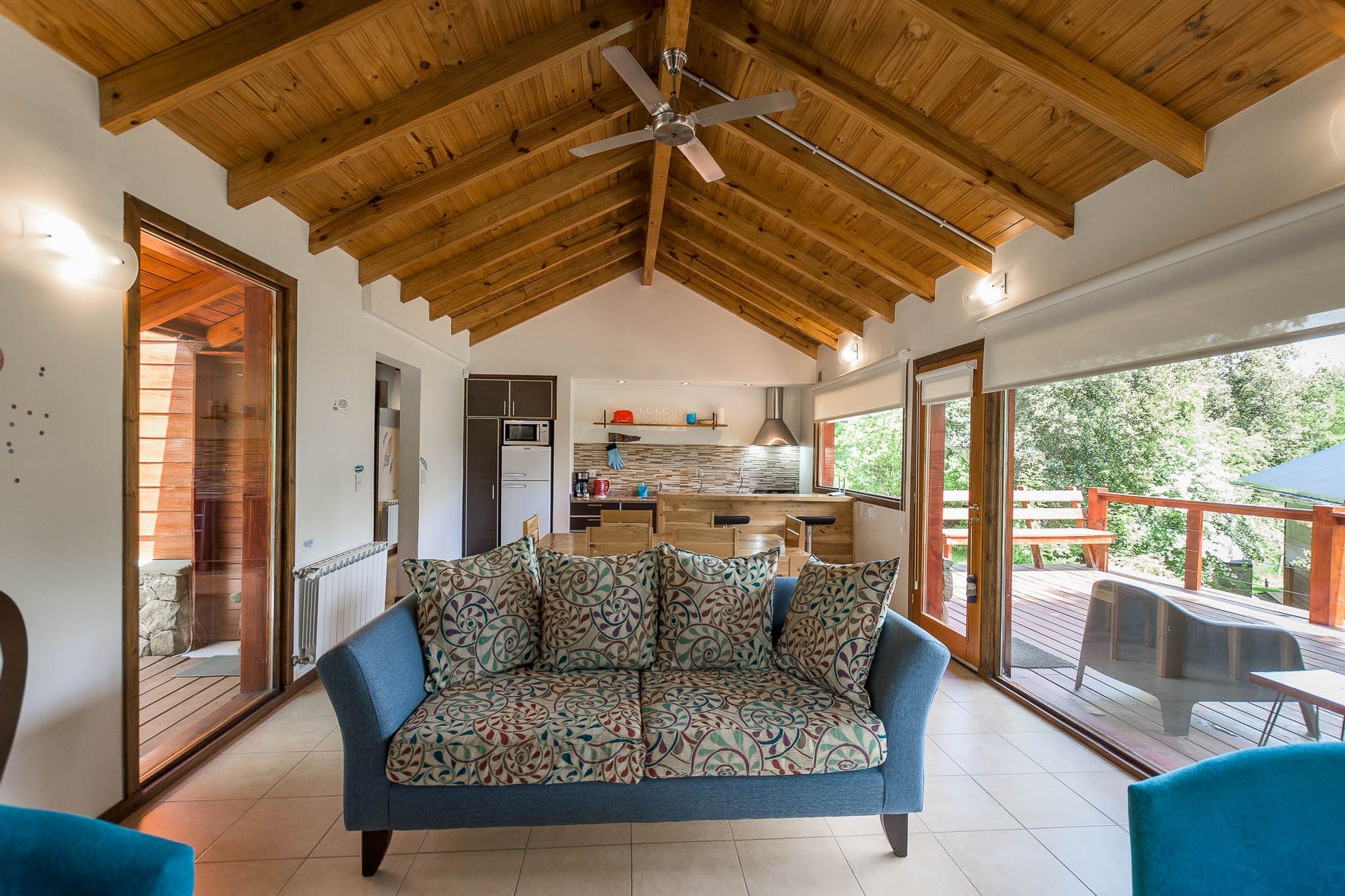 Un luminoso salón con un sofá azul, techo de madera a la vista y grandes puertas de cristal que dan a una terraza y a árboles verdes.