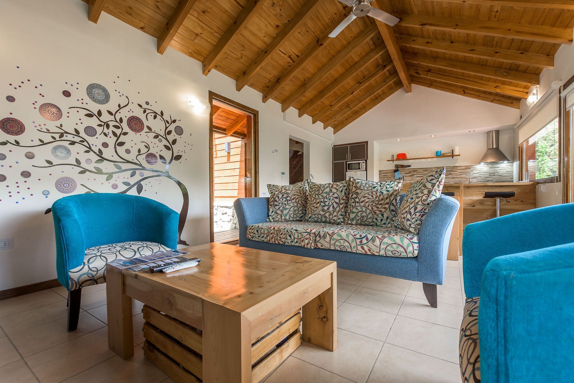 Una acogedora sala de estar con un sofá estampado, dos sillas de color azul brillante, una mesa de centro de madera y un mural pintado con un árbol en la pared.