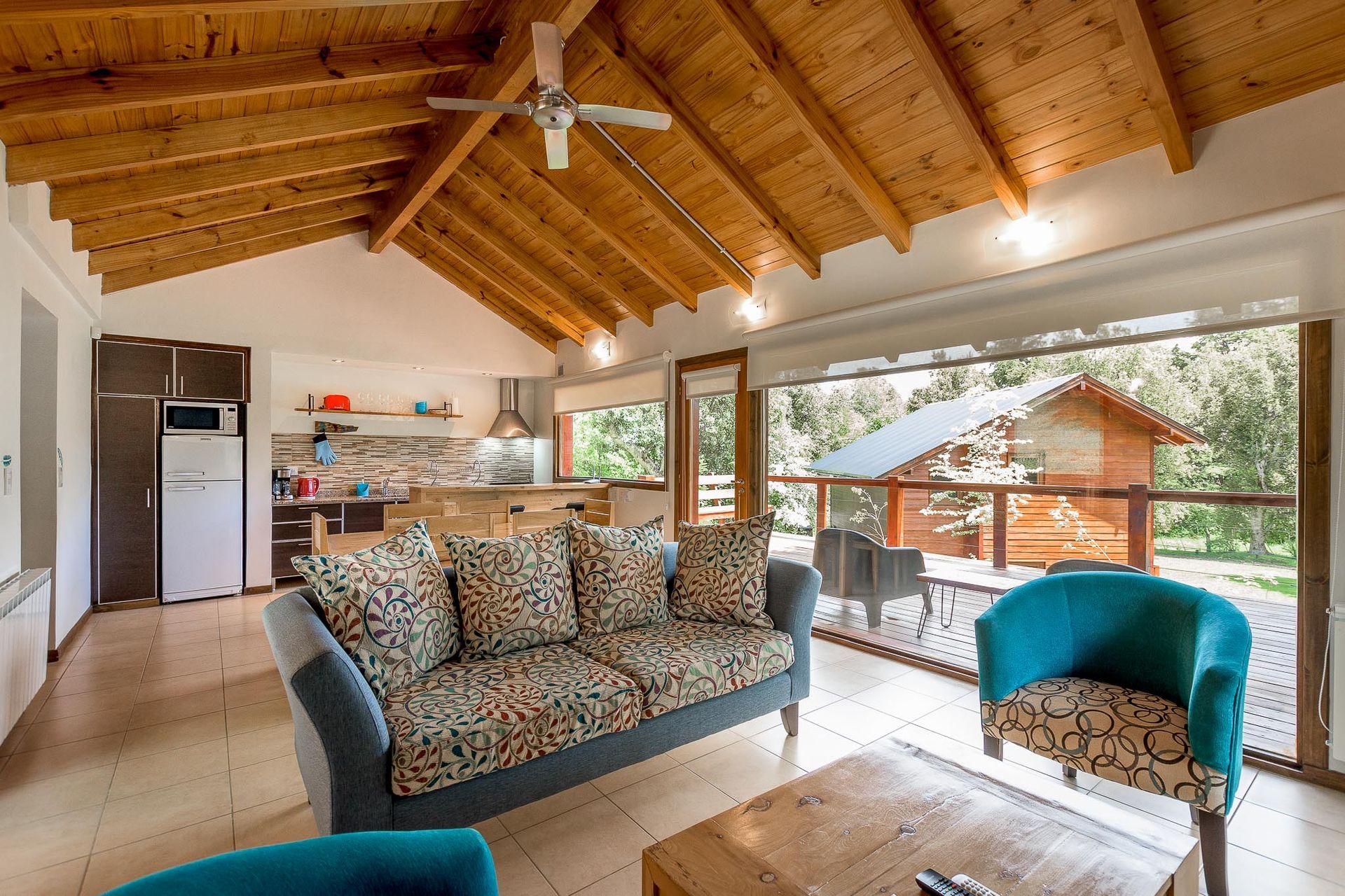 Sala de estar con un sofá estampado, un sillón color turquesa, techo de madera y una zona de cocina visible al fondo.