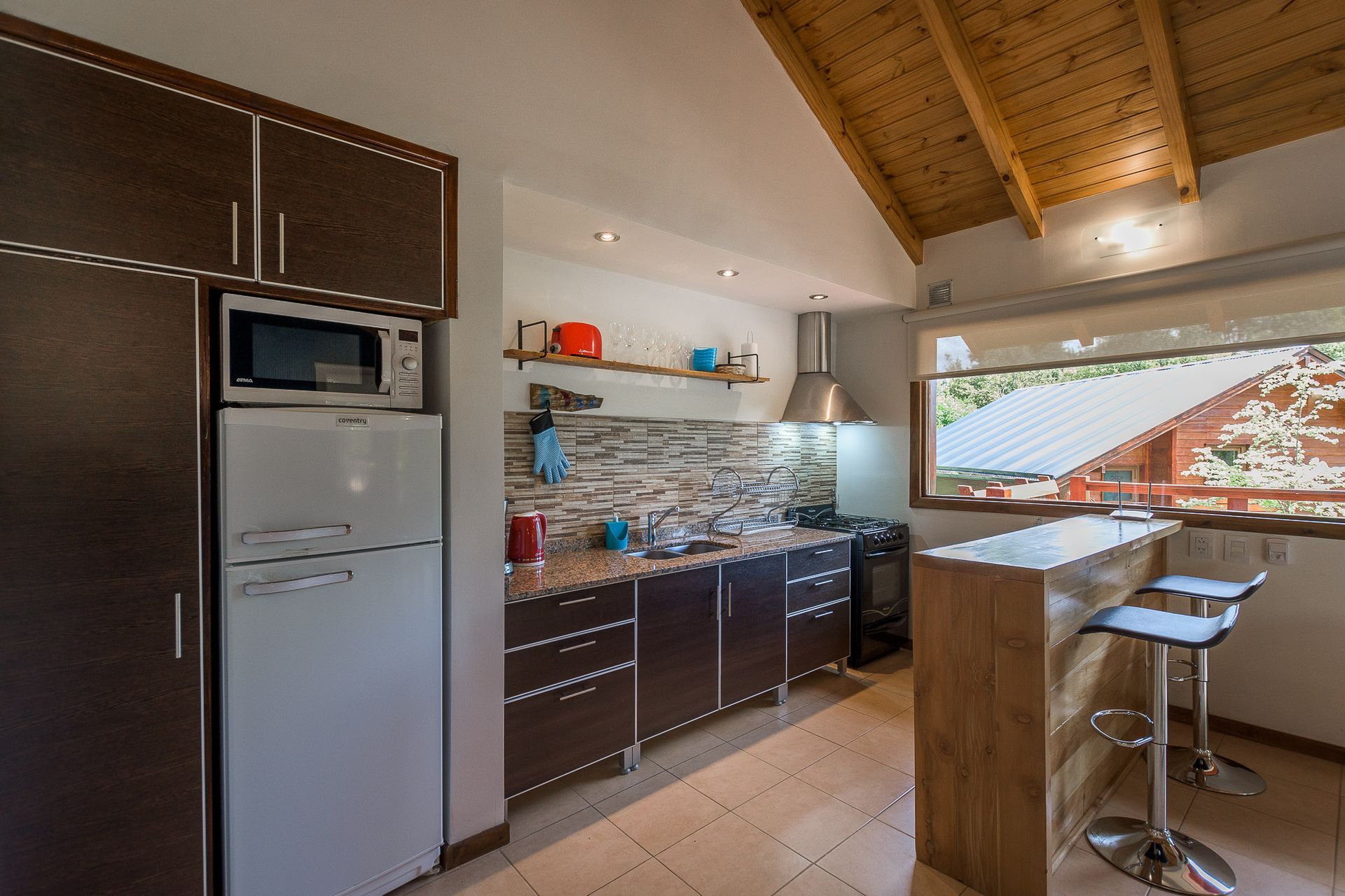 Una cocina con armarios de madera oscura, electrodomésticos blancos, salpicadero de azulejos y una barra de desayuno con dos taburetes.