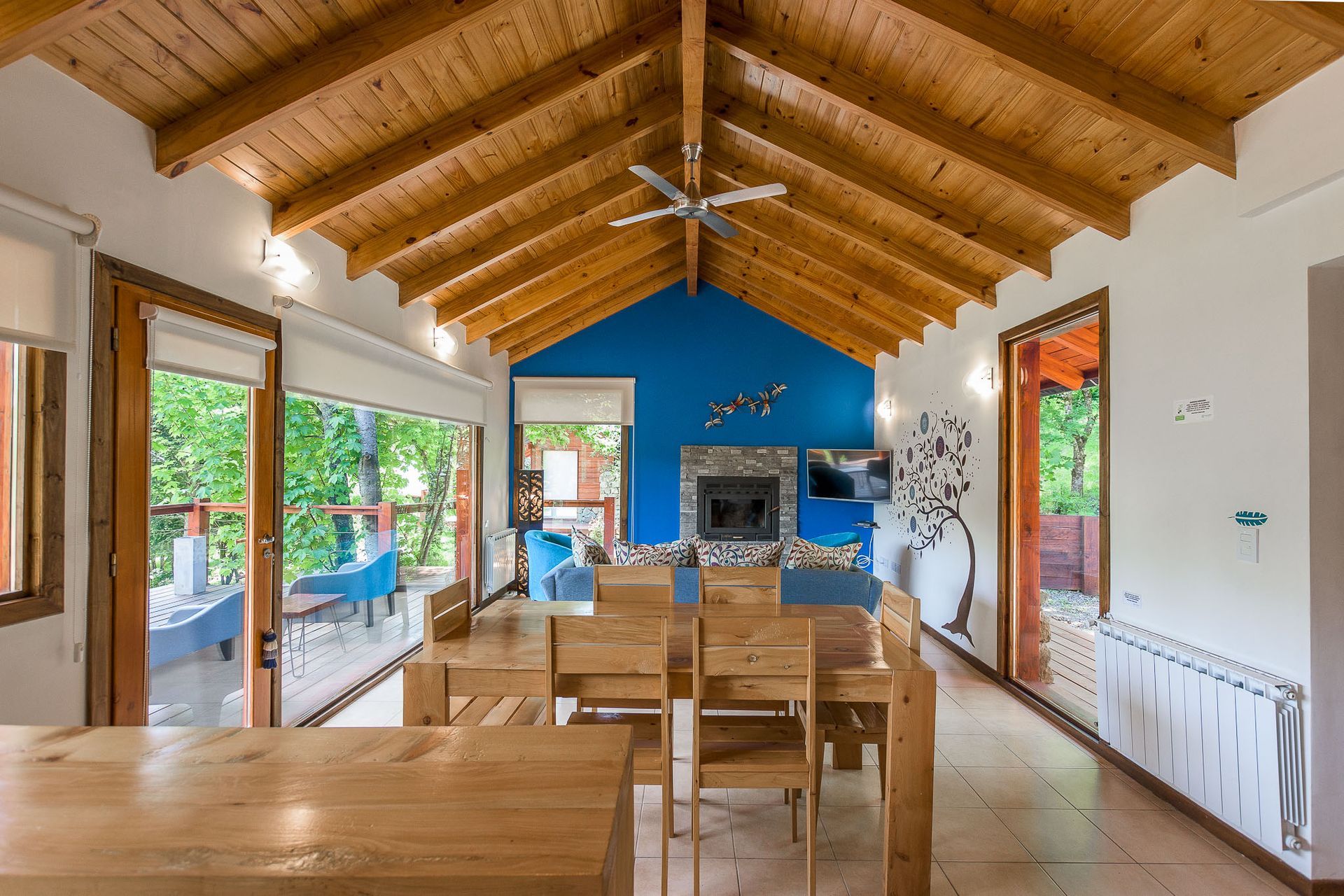 Amplia sala de estar-comedor con techo alto con vigas de madera, muebles de madera y una pared central azul con chimenea.