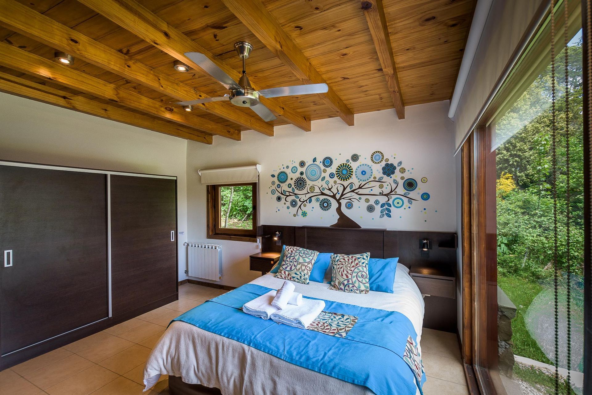 Un dormitorio luminoso con techo de madera, una cama con detalles en azul, un mural de un árbol en la pared y una gran puerta corrediza de cristal.