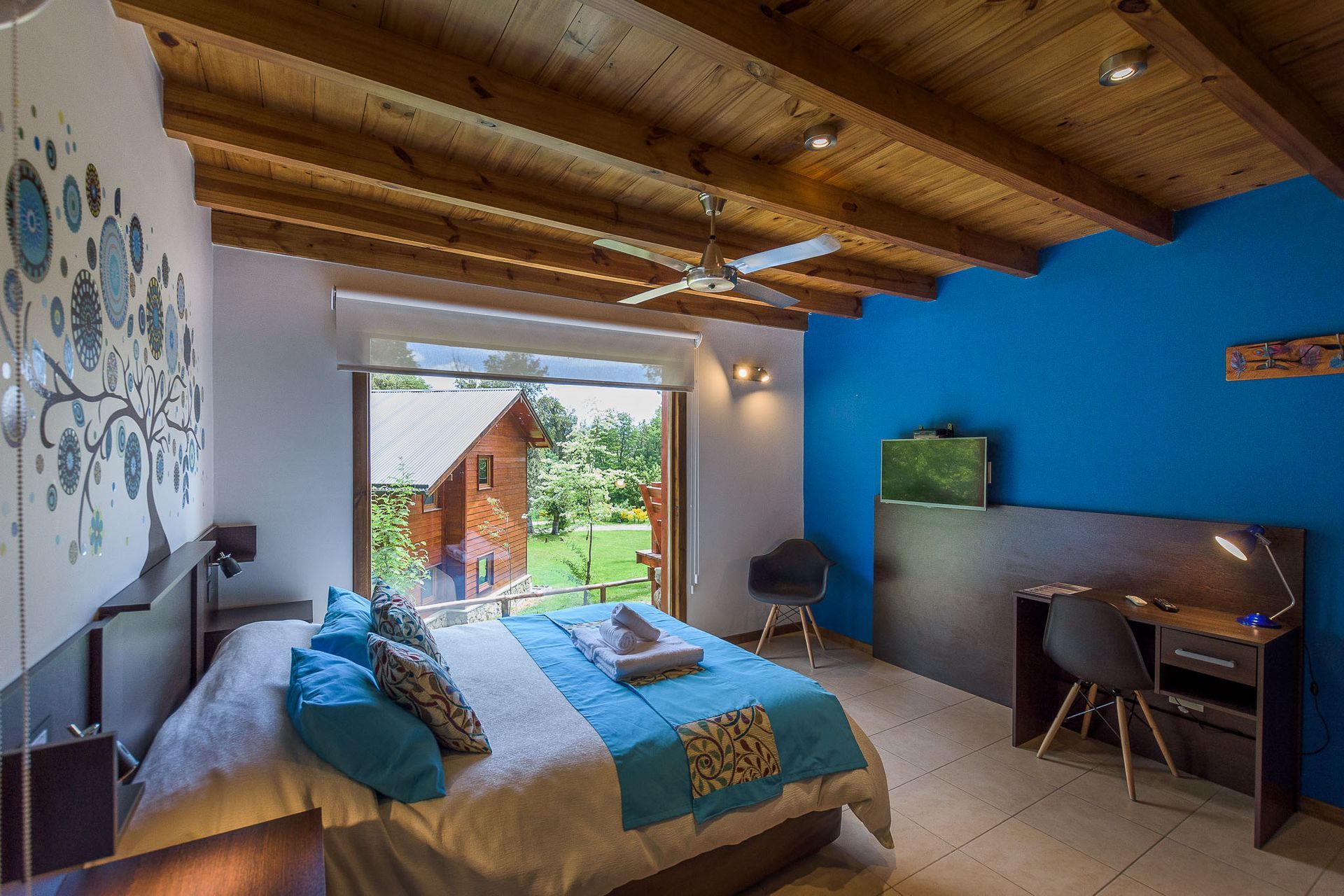 Un dormitorio moderno con techo de madera, una cama con almohadas azules, un escritorio con silla y vistas a un jardín.
