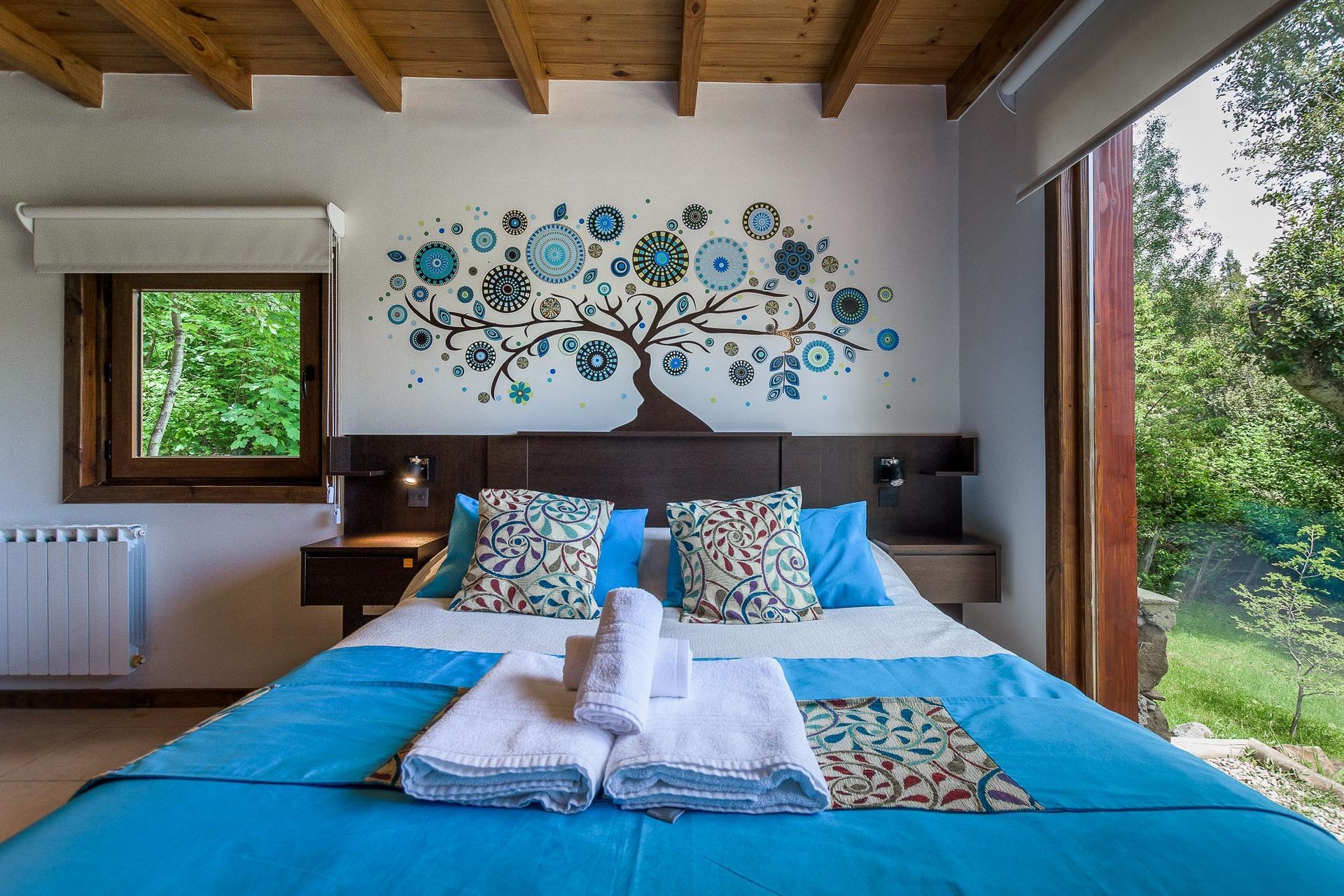 Un acogedor dormitorio cuenta con una cama con una colcha azul brillante y toallas blancas, situada frente a un árbol decorativo en la pared con detalles azules.