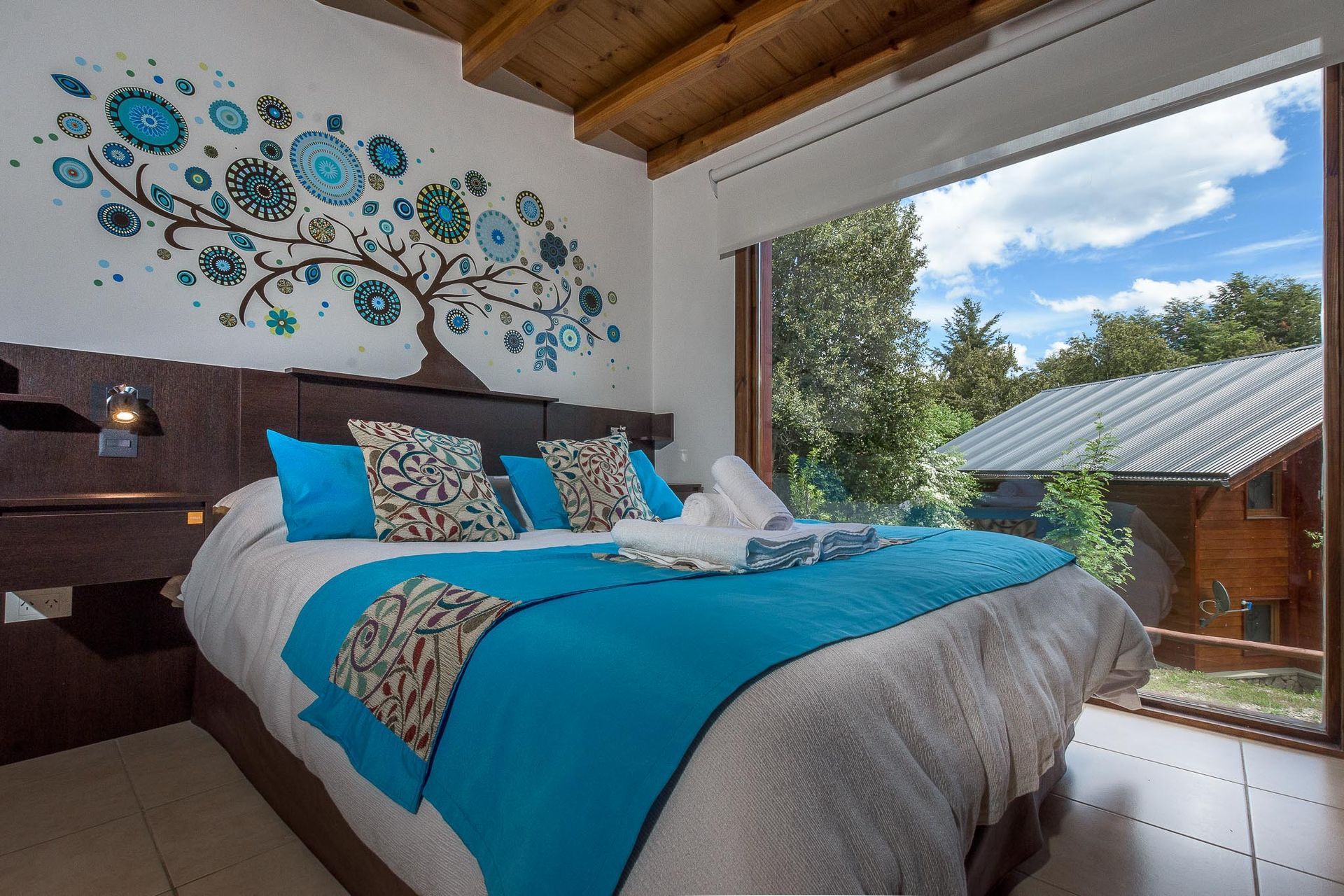 Un dormitorio acogedor con una cama con ropa de cama turquesa y estampada, un mural de pared con motivos de árboles y grandes ventanales.