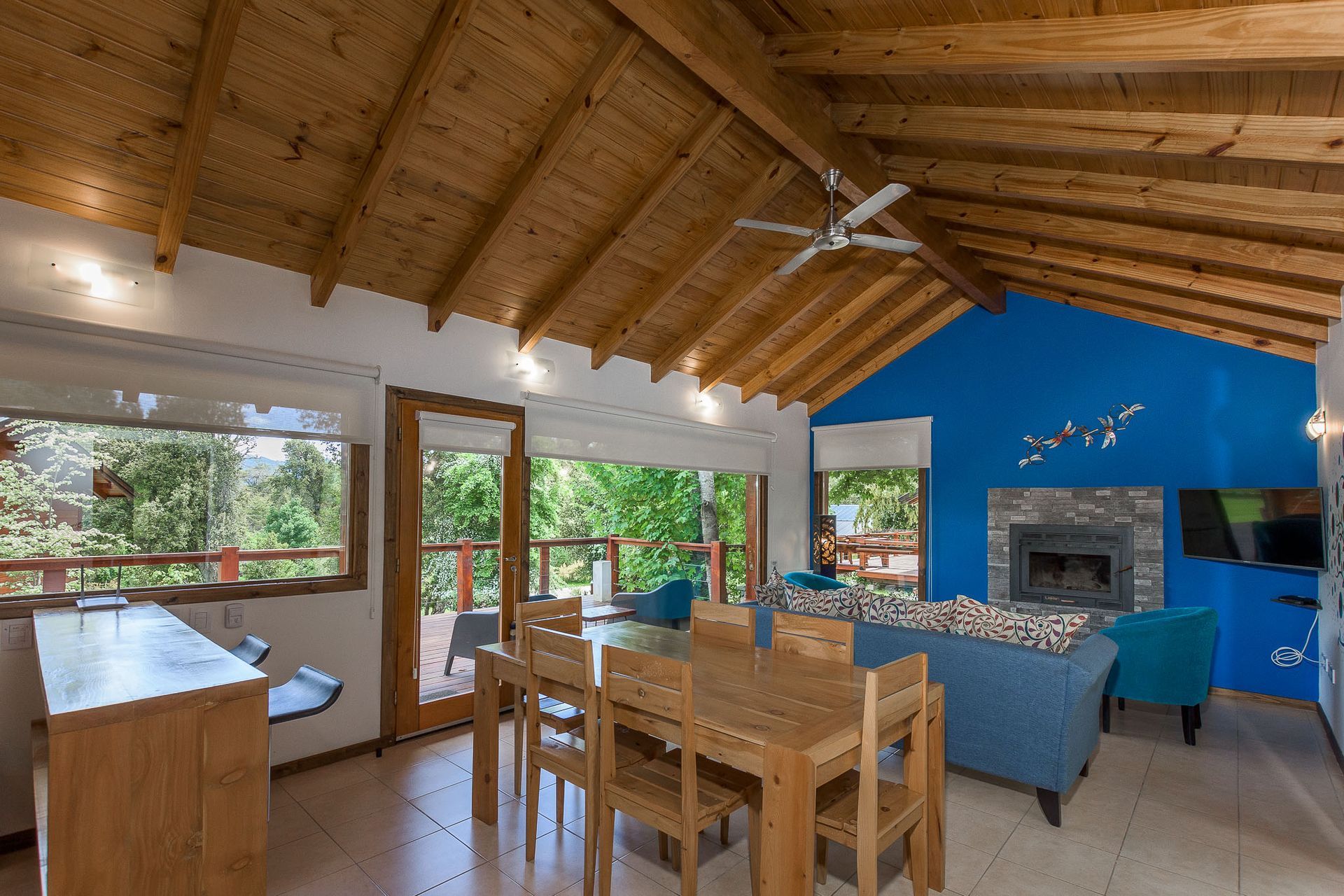 Una habitación luminosa con un techo alto con vigas de madera, una mesa de comedor de madera y una acogedora zona de estar con chimenea y pared azul.