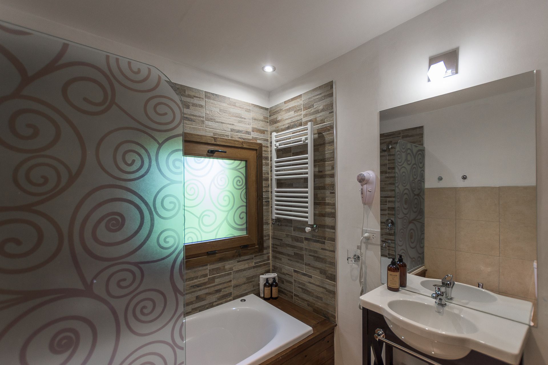 Un baño luminoso con bañera blanca, lavabo con pedestal, toallero eléctrico de pared y biombo estampado para mayor privacidad.