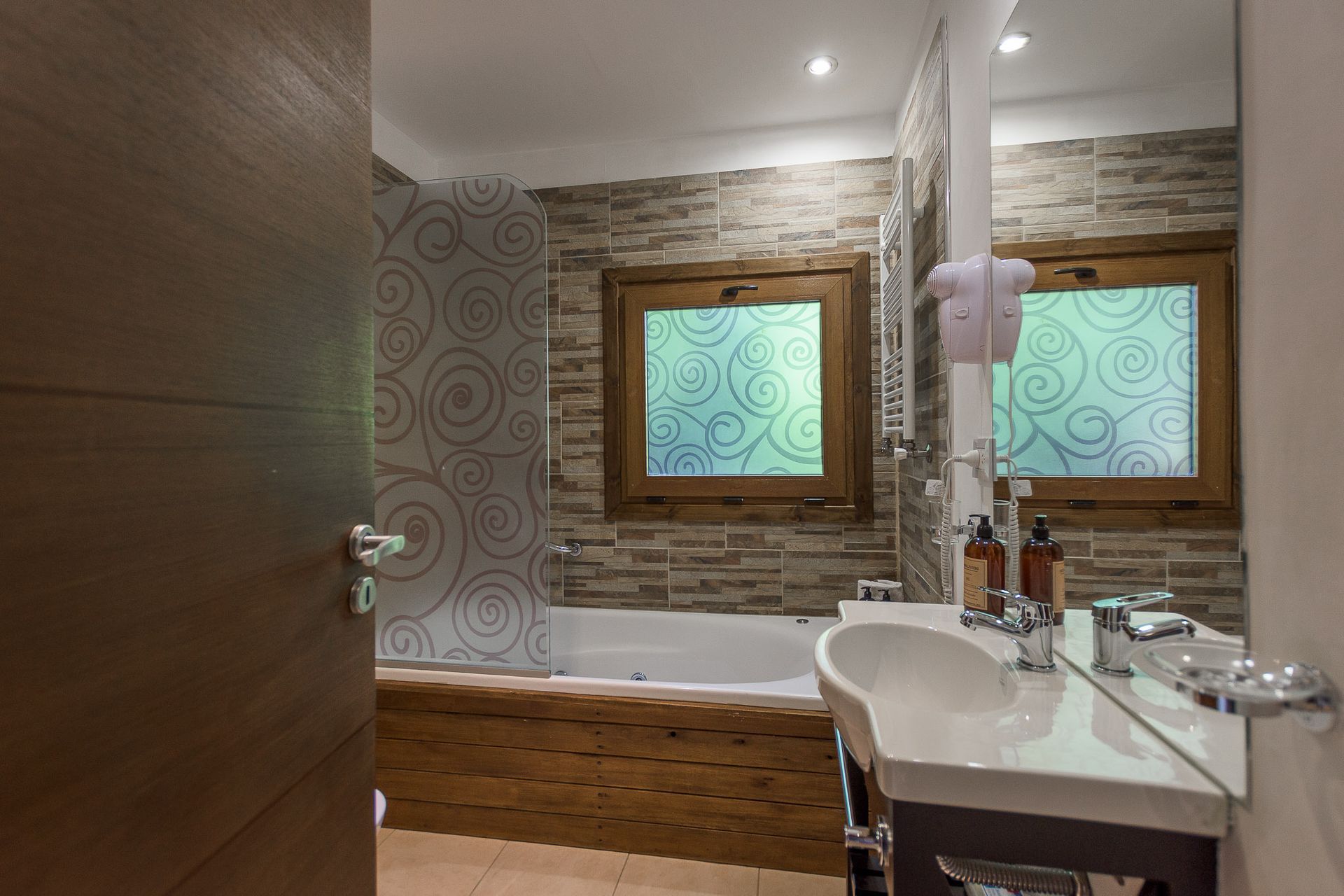 Un baño con bañera, una ventana con marco de madera y cristal esmerilado, y un lavabo blanco sobre encimera, todo ello contra paredes de piedra.