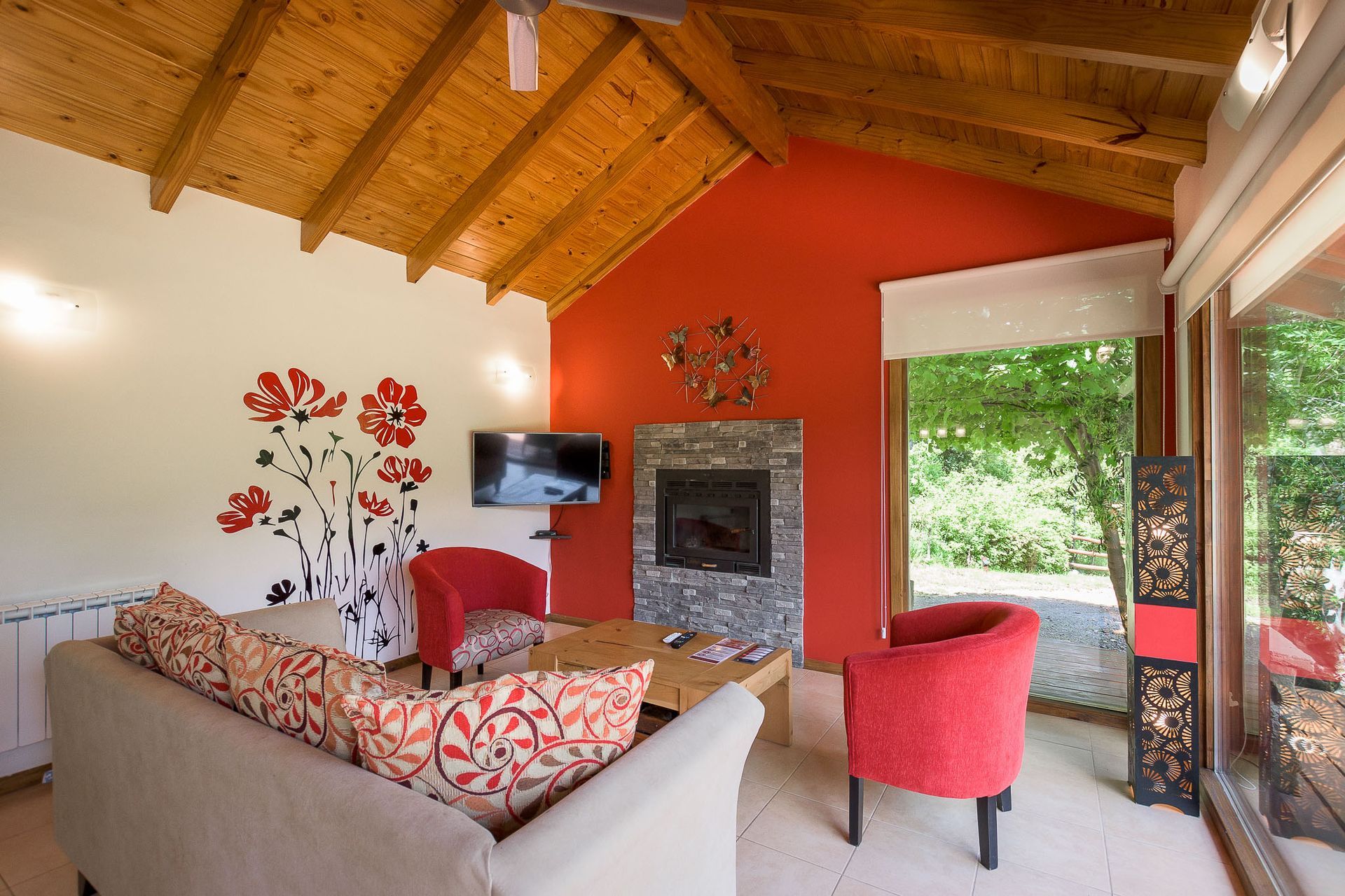 Una acogedora sala de estar con un sofá beige, dos sillones rojos, una chimenea y un cuadro de amapolas bajo un techo con vigas de madera.