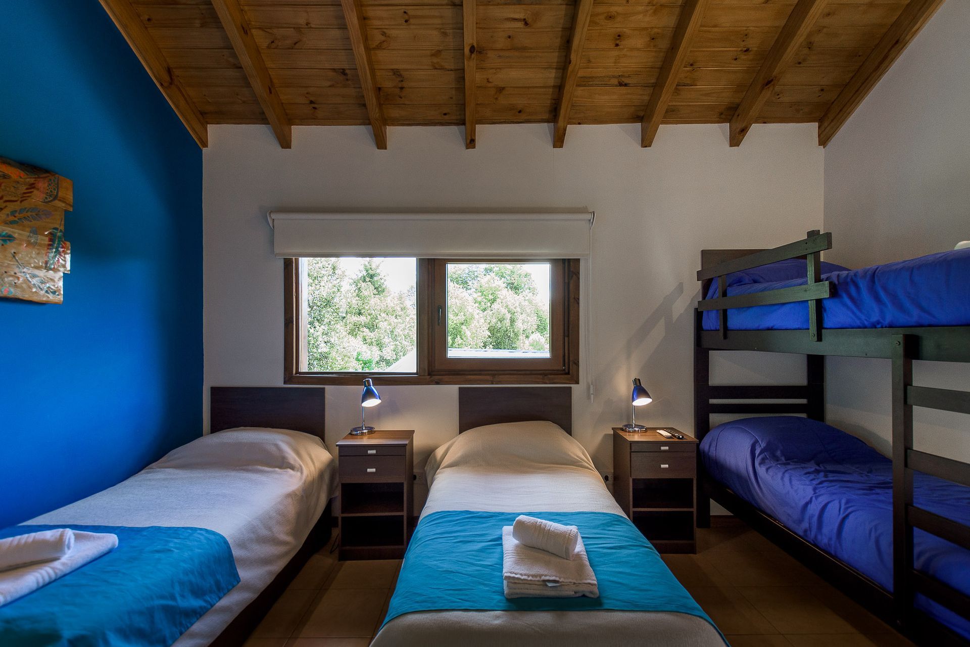 Un dormitorio con una pared azul, un techo inclinado de madera, dos camas individuales y una litera de madera oscura con ropa de cama azul.