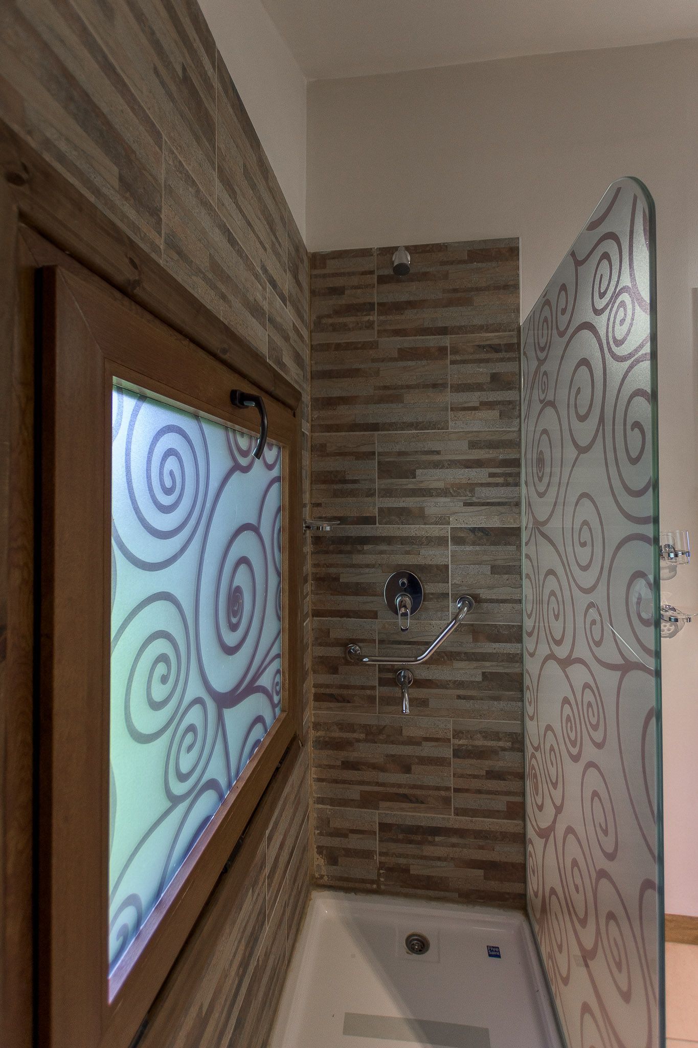 Una cabina de ducha con paredes revestidas de azulejos de piedra apilados, una base blanca y una mampara de cristal con un diseño de espirales blancas.