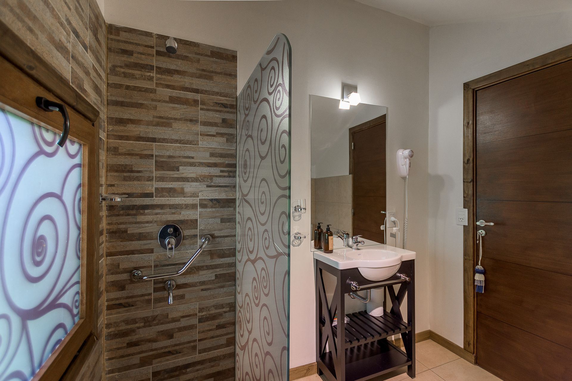 Un baño moderno con ducha a ras de suelo alicatada, mampara de cristal decorada, tocador con lavabo y puerta de madera.