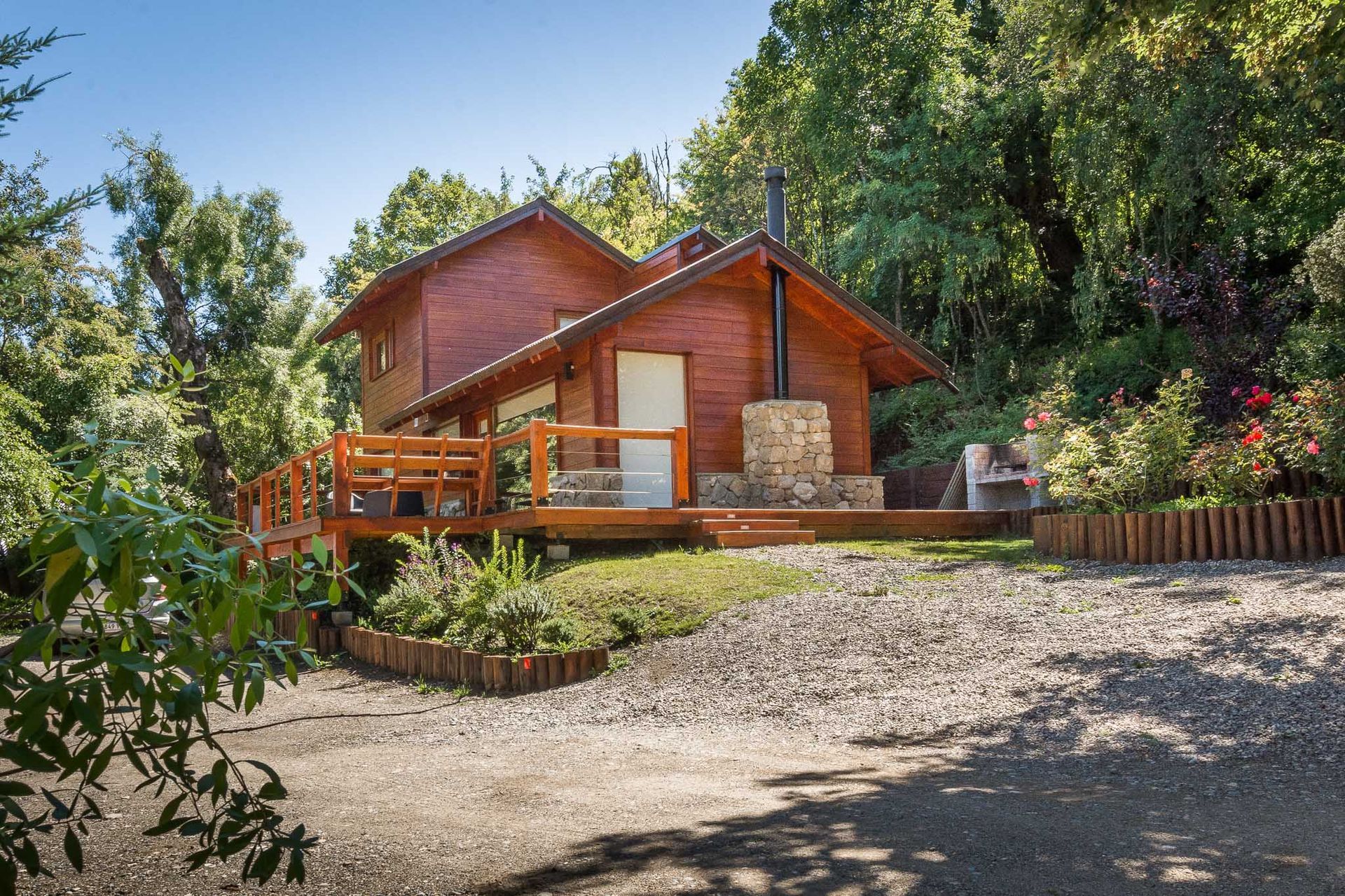 Una pequeña cabaña de madera de dos plantas con chimenea de piedra y terraza que la rodea, situada en un claro soleado del bosque, rodeado de árboles.