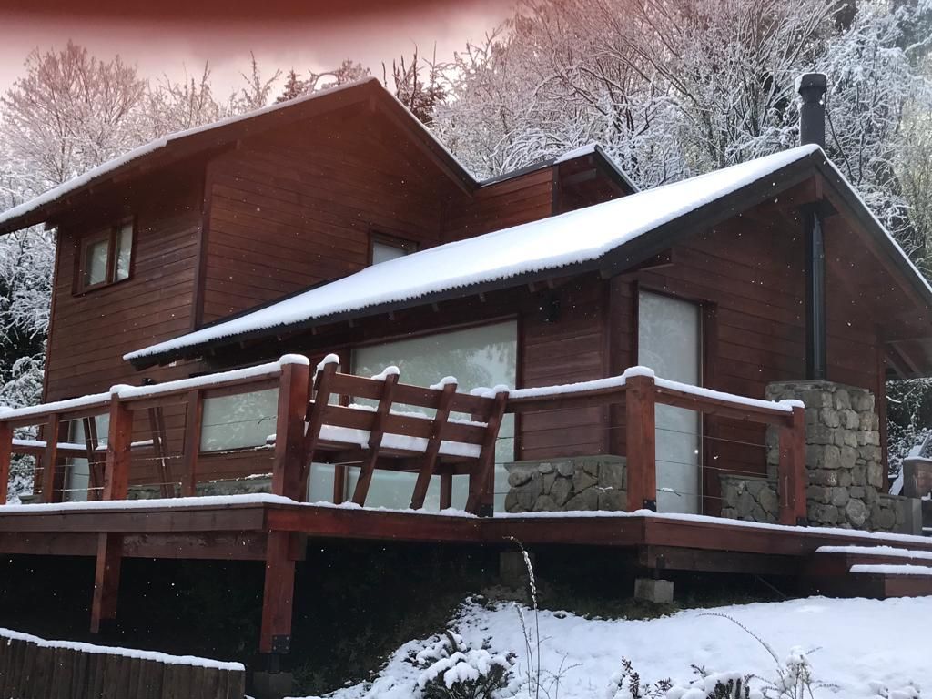 Una cabaña de troncos marrones de dos pisos con terraza de madera y chimenea de piedra, situada en un paisaje nevado.