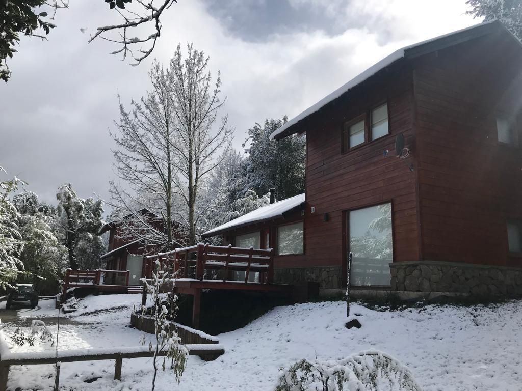 Una cabaña de dos plantas, revestida de madera y con terraza, rodeada por una ligera capa de nieve en un día nublado.