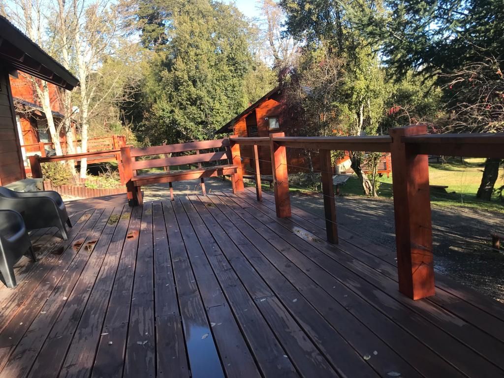 Una terraza de madera con barandilla y banco, con vistas a una zona soleada y boscosa, con otros edificios de madera en la distancia.