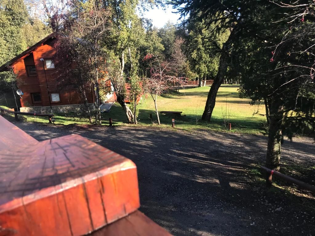 Vista desde una terraza de madera con vistas a un sendero de grava, una cabaña rústica de troncos y un jardín con árboles al atardecer.