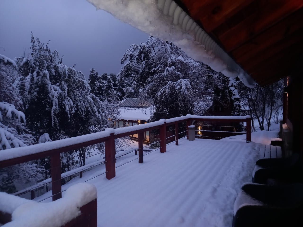 Un balcón nevado al atardecer, con árboles cubiertos de nieve y una pequeña cabaña iluminada al fondo.