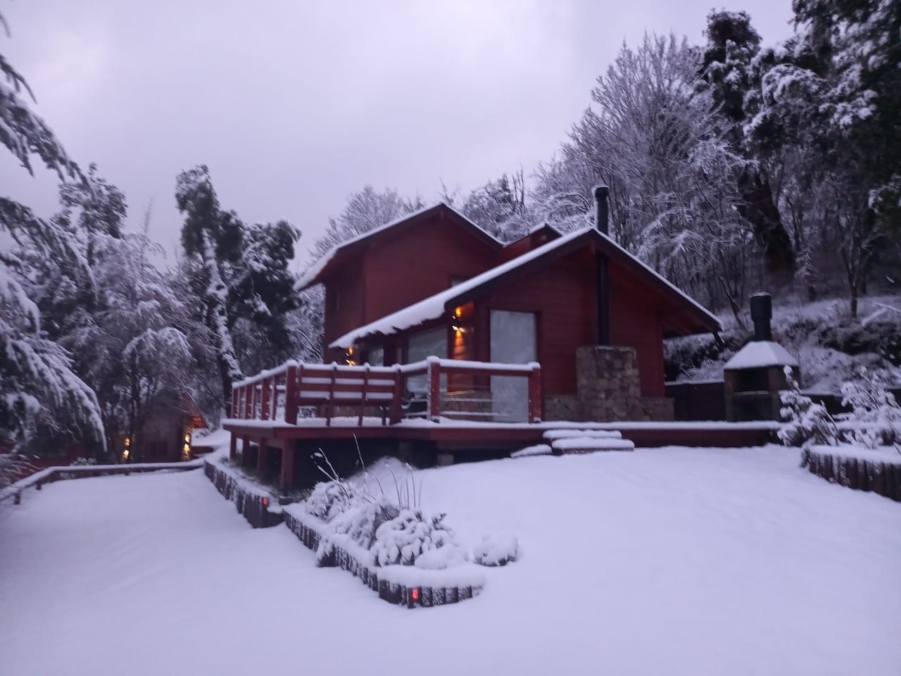 En invierno, una cabaña de madera de color marrón rojizo, con terraza y chimenea de piedra, se alza en un paisaje boscoso y nevado.