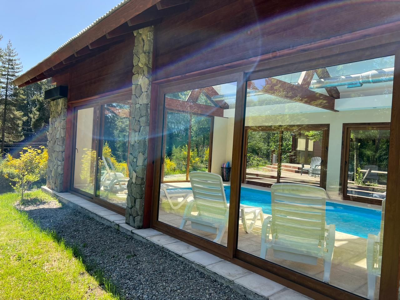 Una piscina bajo una estructura acristalada con tumbonas blancas, visible a través de ventanales que van del suelo al techo.