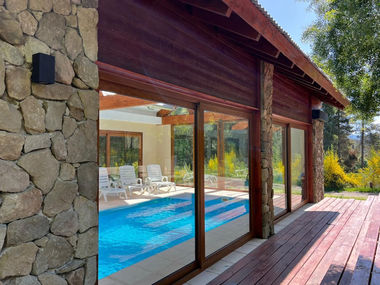 Una piscina vista a través de puertas corredizas de vidrio desde una terraza de madera, bordeada por un muro de piedra rústica y revestimiento de madera.