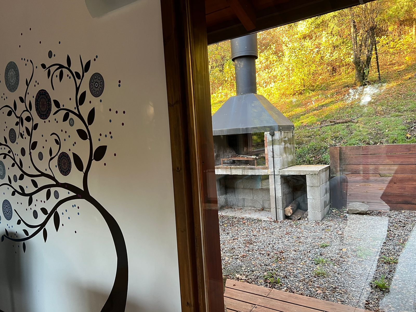 Vista desde el interior hacia una parrilla de piedra al aire libre en un patio arbolado cubierto de grava con un vinilo decorativo de un árbol en la pared.
