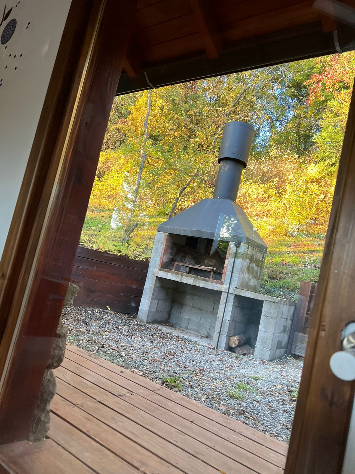 Una chimenea de piedra al aire libre, vista desde una terraza de madera, con un telón de fondo de árboles otoñales de hojas amarillas.