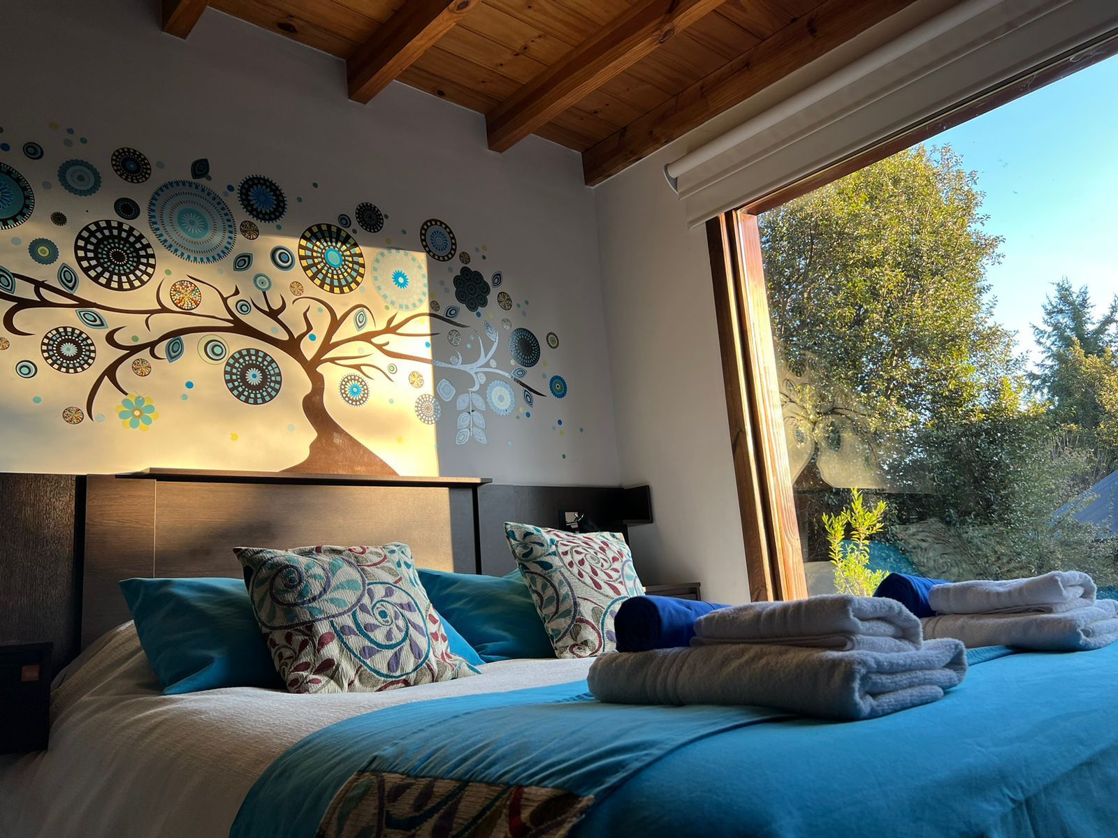 Un dormitorio con una cama tamaño king, ropa de cama color turquesa y un gran mural de un árbol en la pared, iluminado por la luz del sol que entra por una amplia ventana.