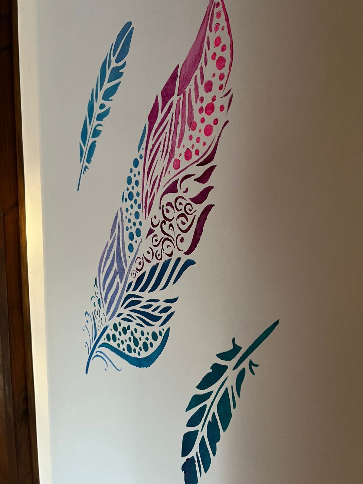 Un mural de pared con una plantilla de pluma grande, ornamentada y multicolor, flanqueada por dos plantillas de plumas más pequeñas de color verde azulado liso.