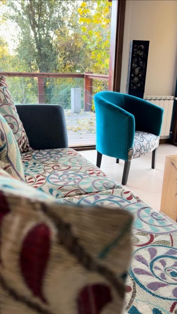 En la sala de estar hay un sofá estampado, con una silla auxiliar de color turquesa cerca y una puerta de cristal con vistas a los árboles.