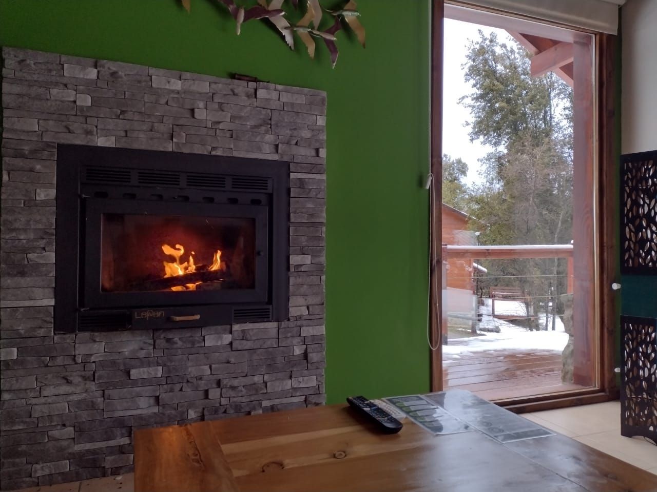 Una acogedora sala de estar con chimenea revestida de piedra y fuego, junto a una ventana con vistas a un patio nevado y a los árboles.
