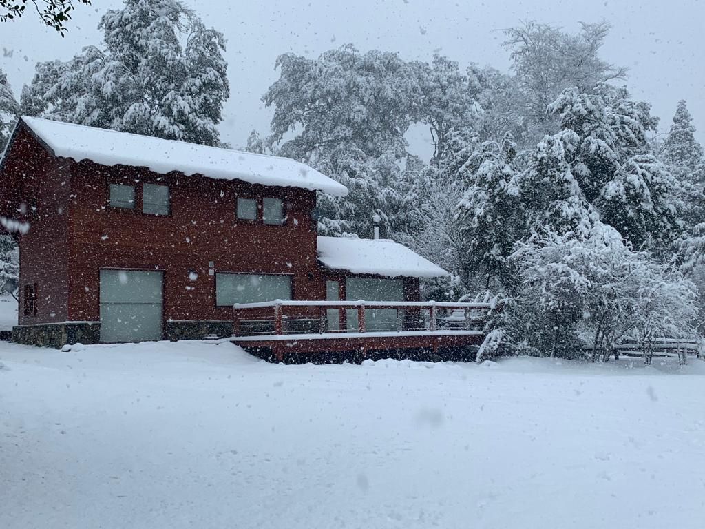 Una cabaña marrón de dos pisos se alza en un paisaje nevado durante una ligera nevada, rodeada de árboles de hoja perenne cubiertos de nieve.