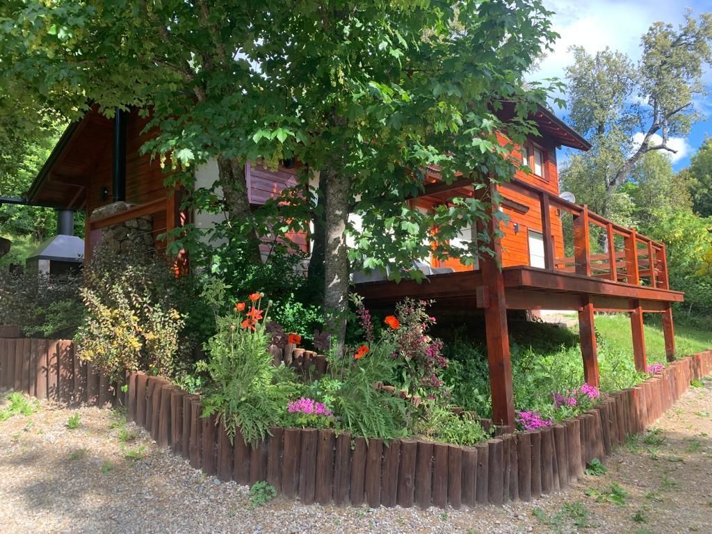 Una cabaña rústica de madera con terraza, parcialmente sombreada por grandes árboles y rodeada por un macizo de flores con un borde de troncos.