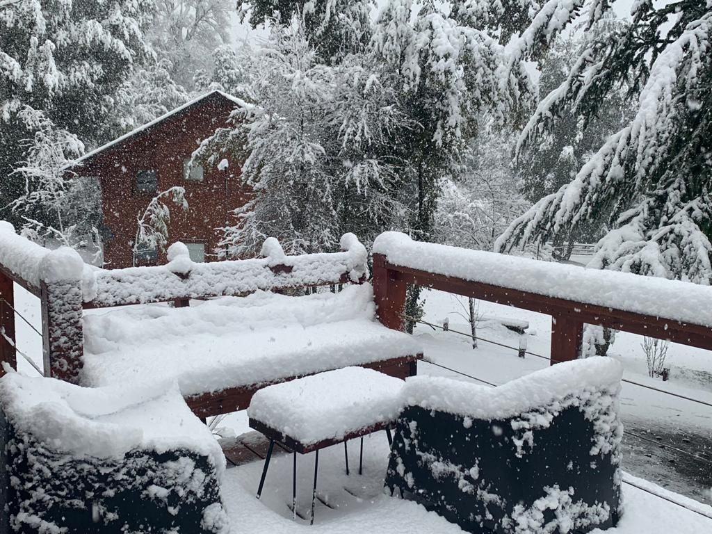Una terraza de madera con muebles de exterior completamente cubierta de nieve blanca y fresca, con vistas a un paisaje boscoso y nevado.