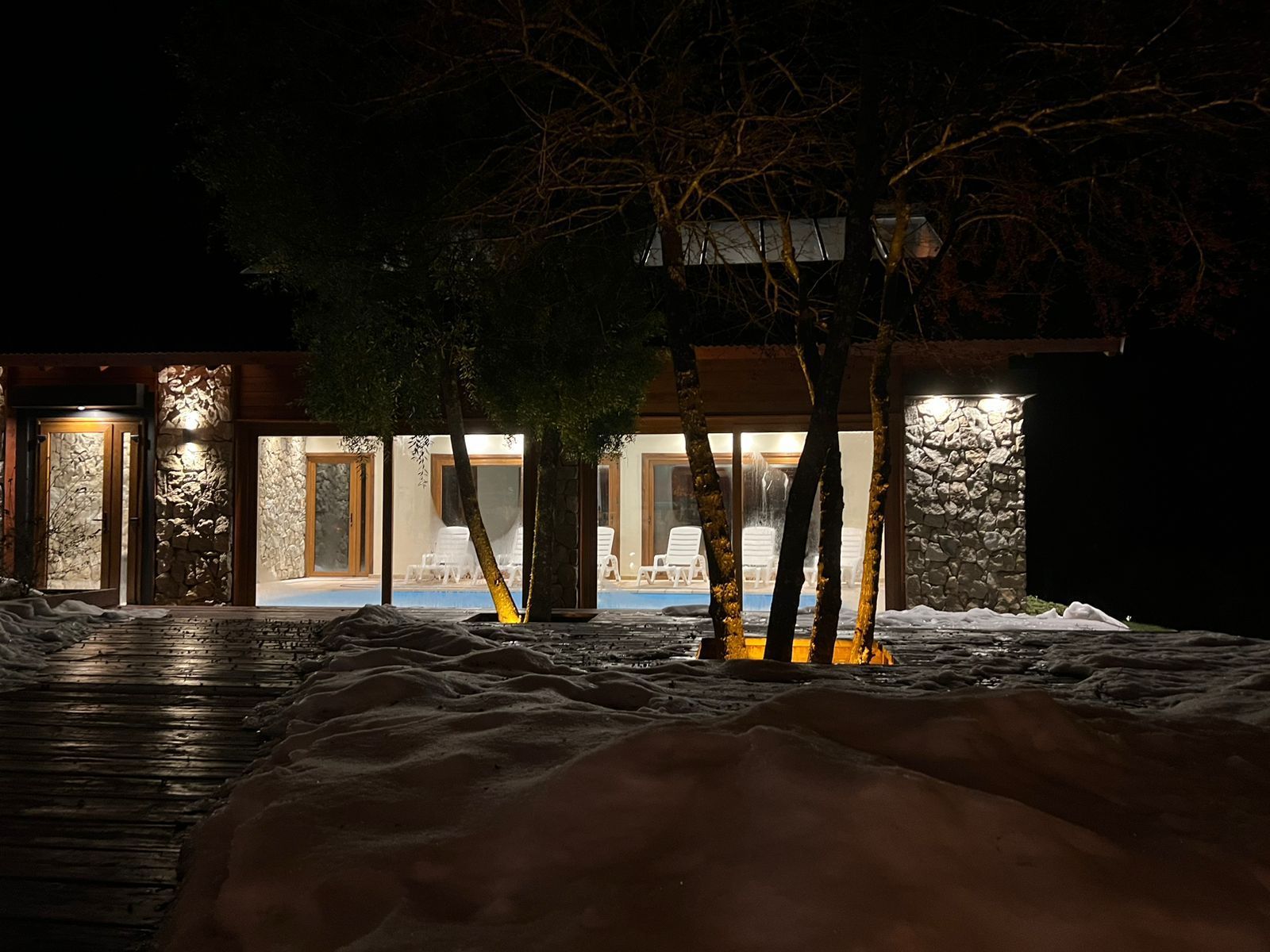 Una piscina cubierta con tumbonas blancas, visible de noche a través de grandes paredes de cristal, enmarcada por árboles y suelo cubierto de nieve.