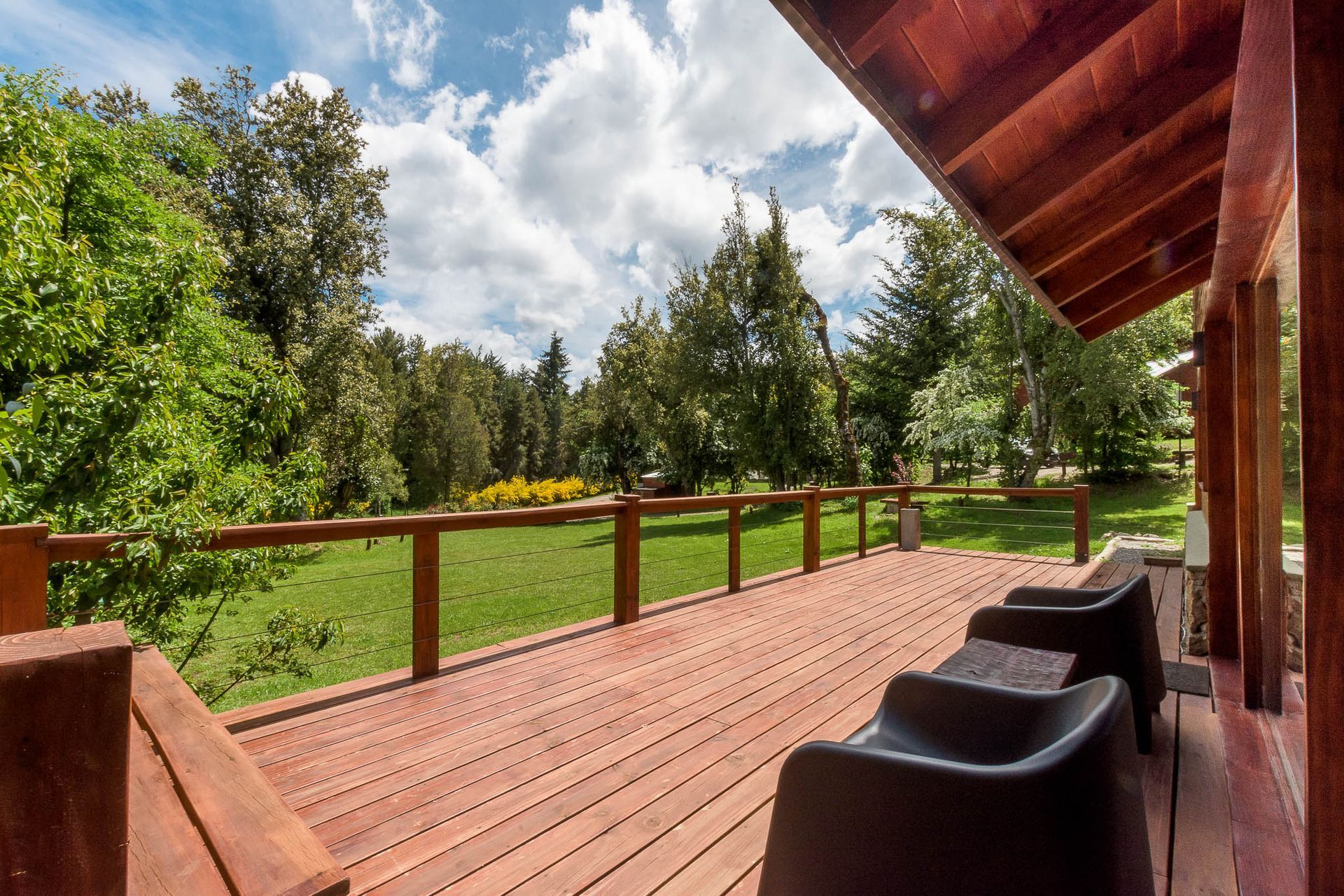 Una terraza de madera con dos sillas oscuras con vistas a un césped verde y frondoso y a los árboles bajo un cielo nublado.