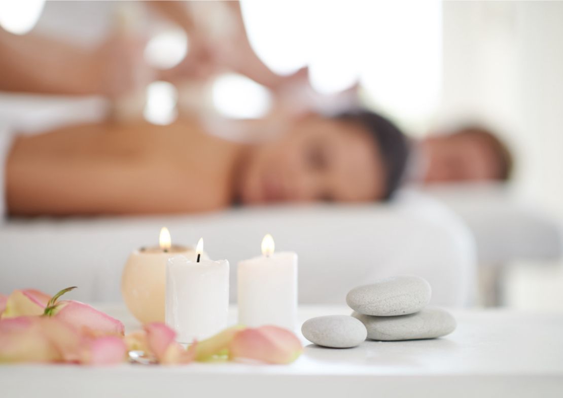 Un tratamiento de spa con personas recibiendo masajes desenfocadas al fondo y velas y piedras en primer plano.