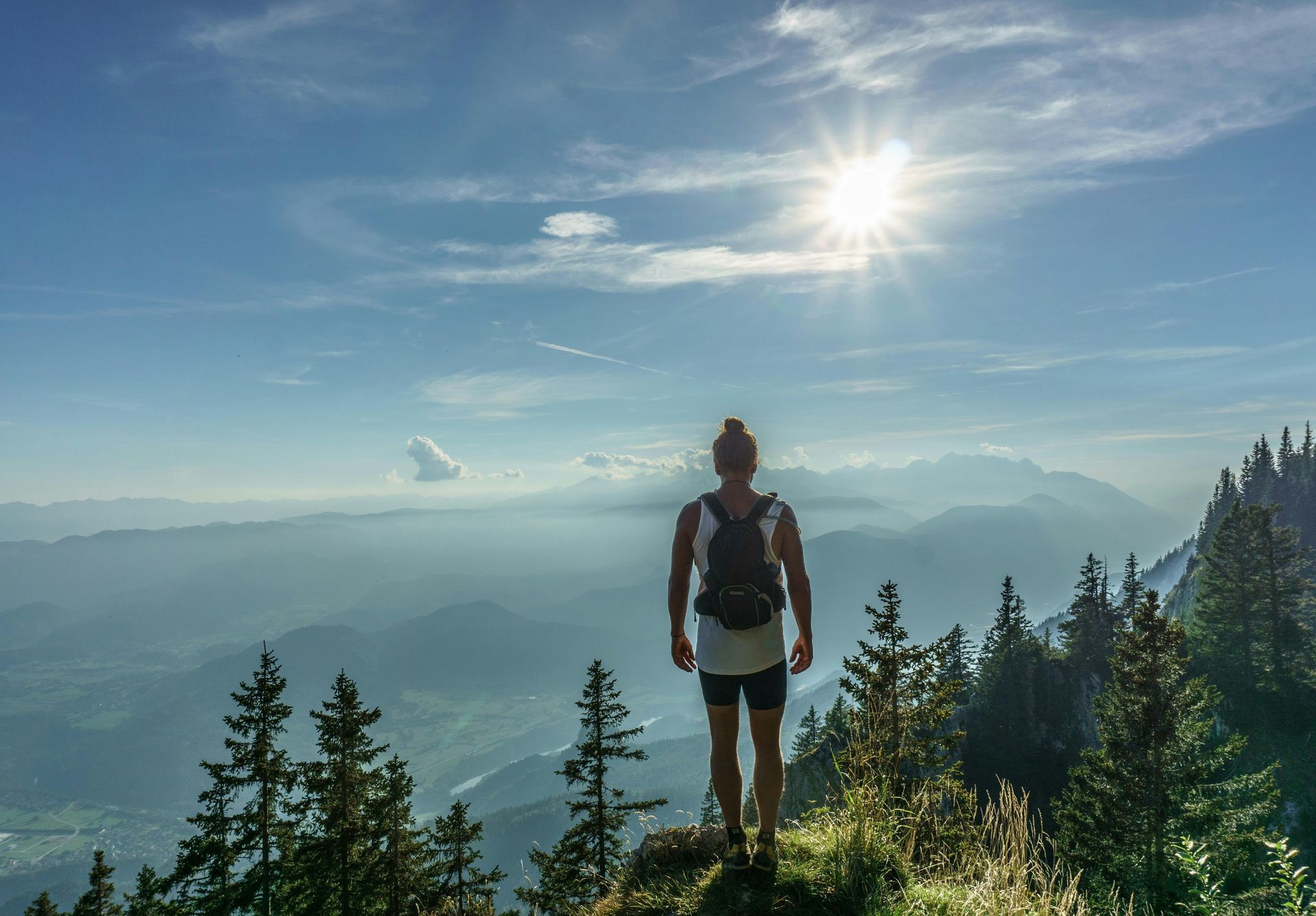 Una persona con una mochila está de pie en la cresta de una montaña, contemplando un valle y laderas boscosas bajo un sol brillante.