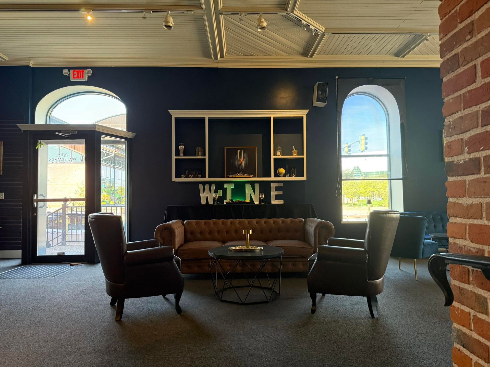 Cozy lounge with a tan sofa, dark chairs, round table, and large window, accented by a “WINE” sign.