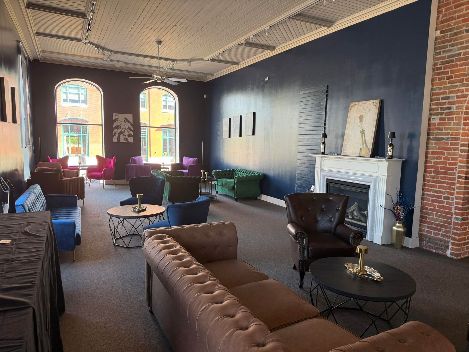 Stylish lounge with tufted sofas, round tables, a fireplace, and large arched windows in a brick-walled room