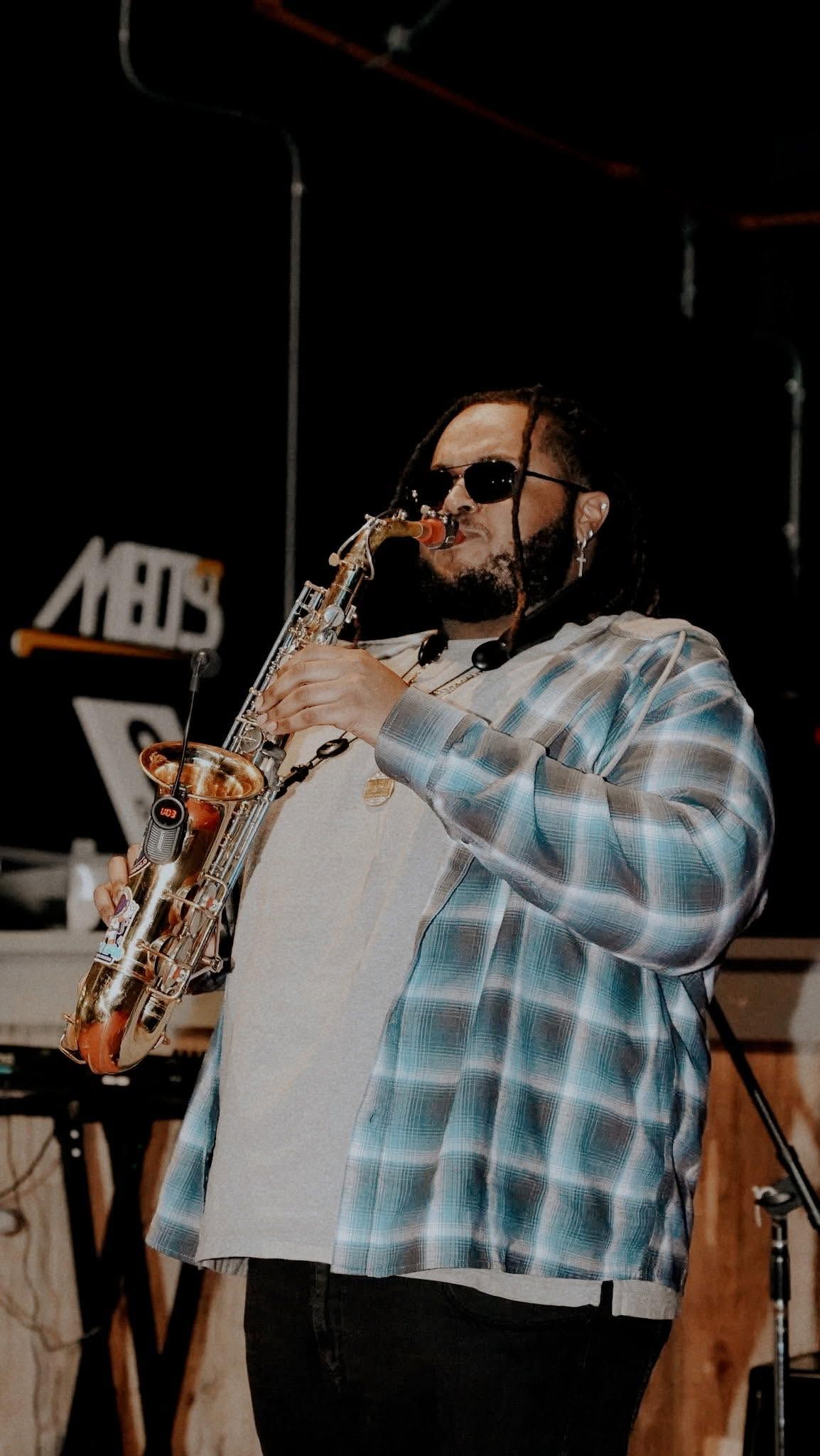 Man playing saxophone on stage in a blue plaid shirt and sunglasses