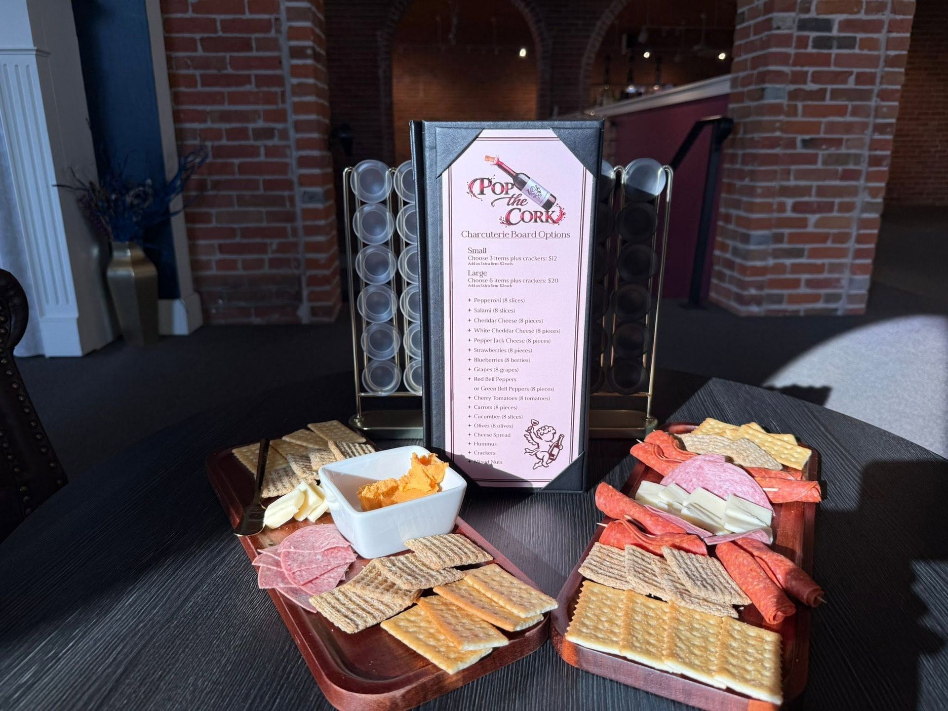Catered buffet table with wrapped sandwiches, chips, and dip beside a menu sign in a brick venue