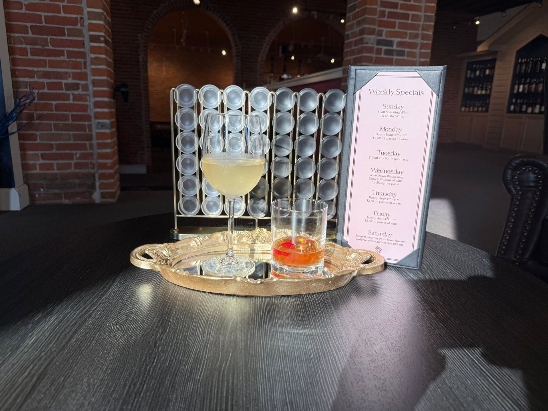 Cocktail tray with a pale drink, orange garnish, and menu card on a black table in a brick interior.