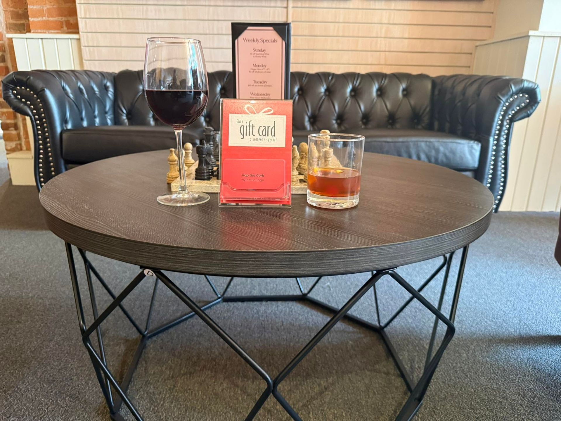 Round coffee table with two drinks, cigars, and bottles in a lounge with a black leather couch.