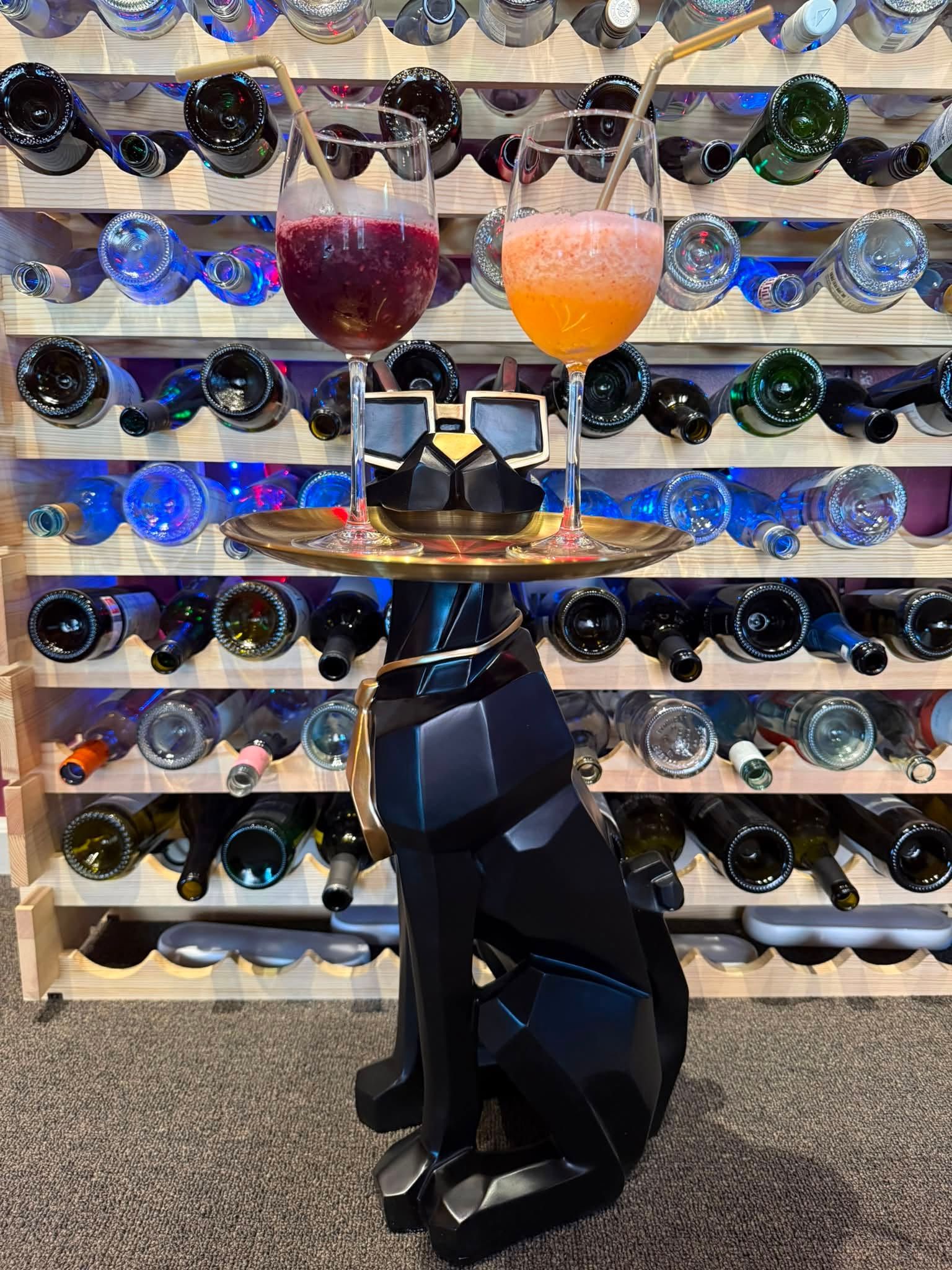 Black cat sculpture on a carpeted floor in front of a wine rack, with two colorful drinks on a stand.