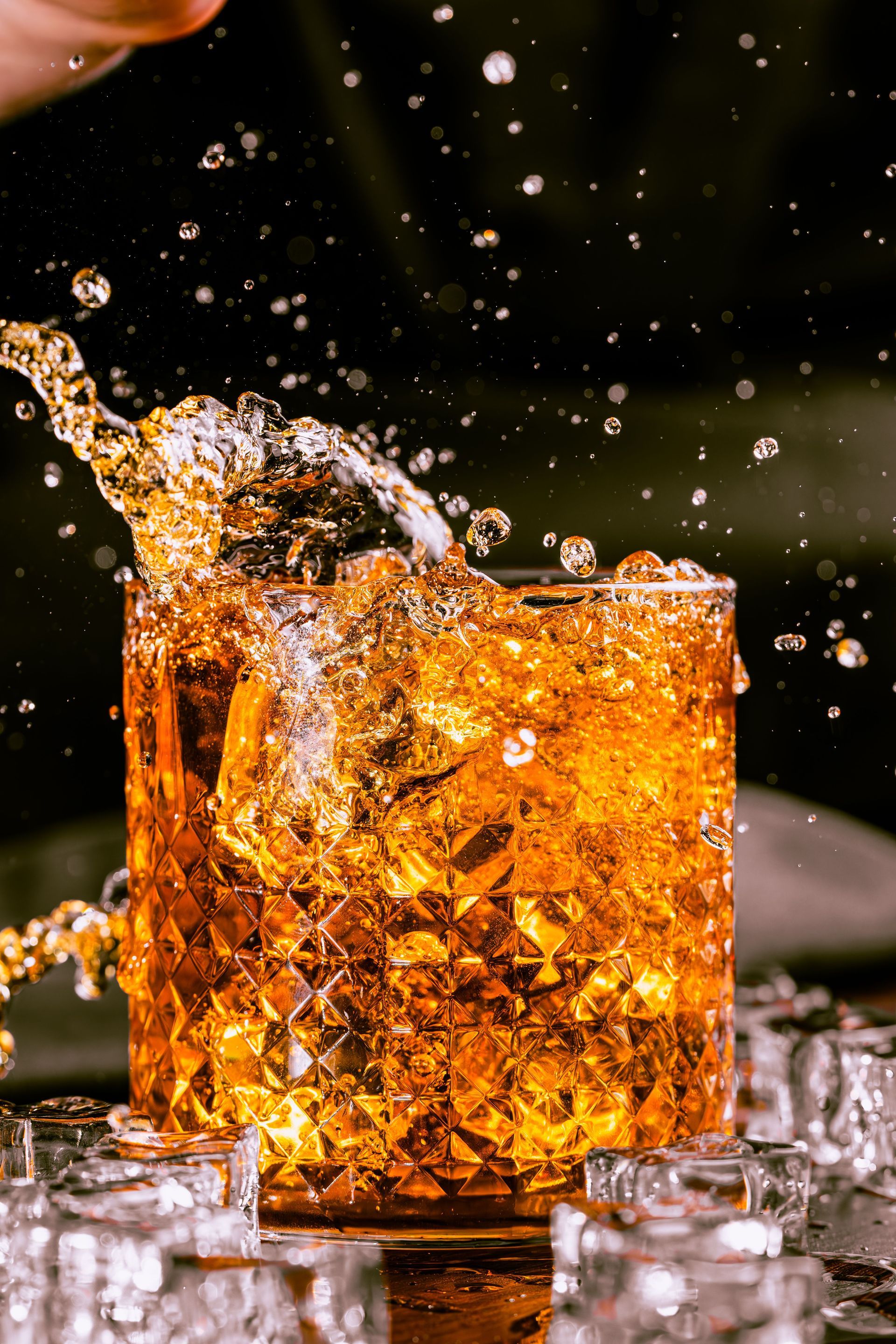 A whiskey glass with amber liquid splashing over ice against a dark background