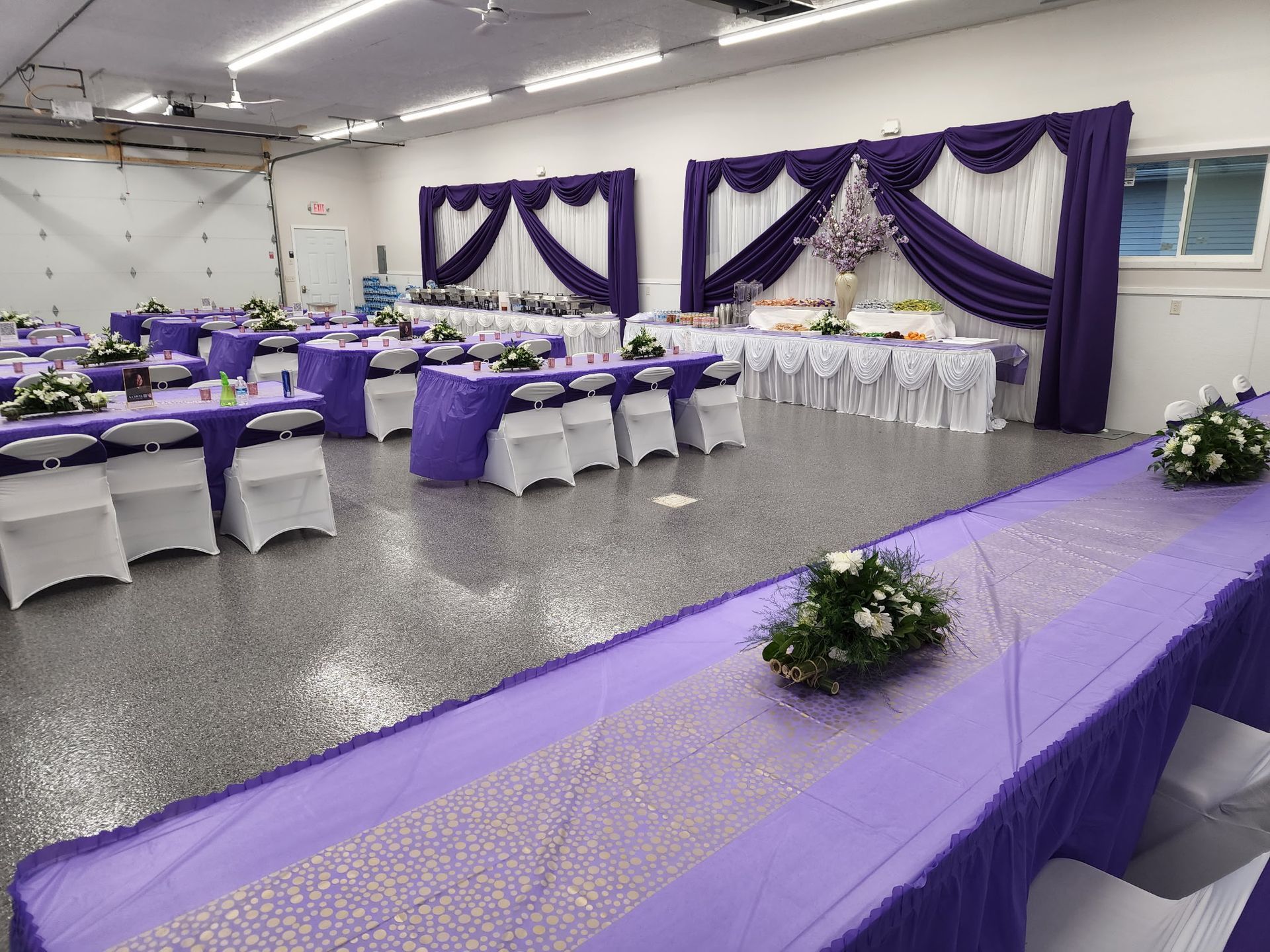 Decorated banquet hall with purple and white table settings and draped stage backdrop